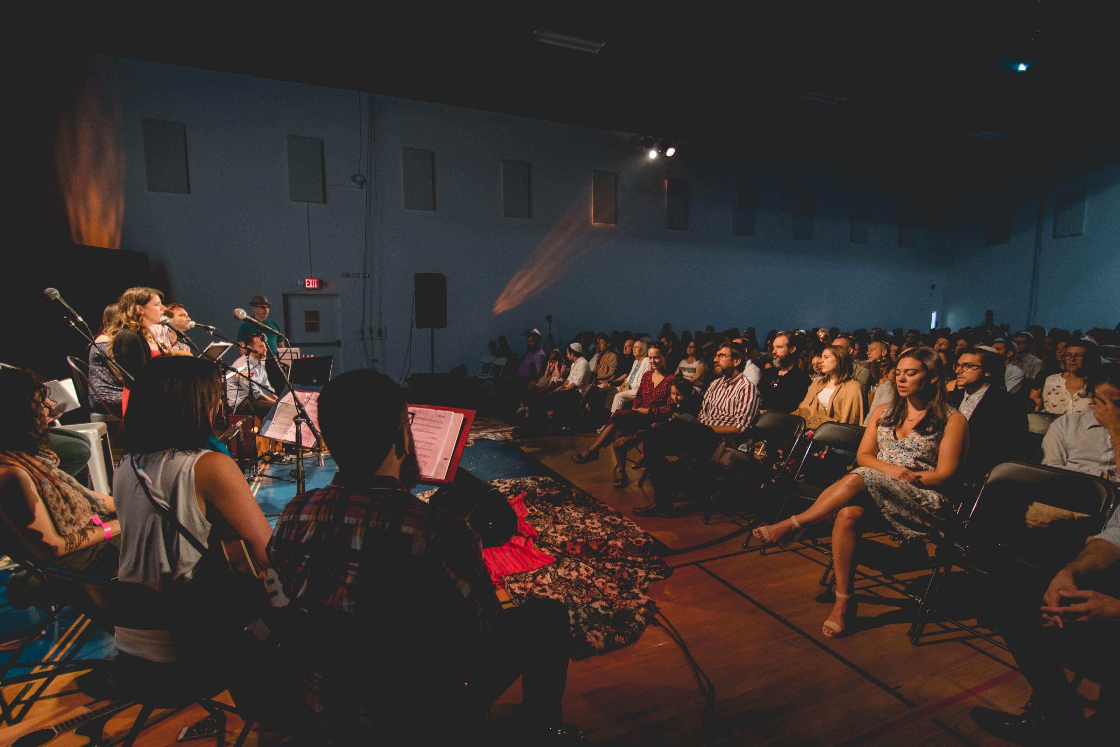 A small audience listens to a performance on stage in a dimly lit auditorium.