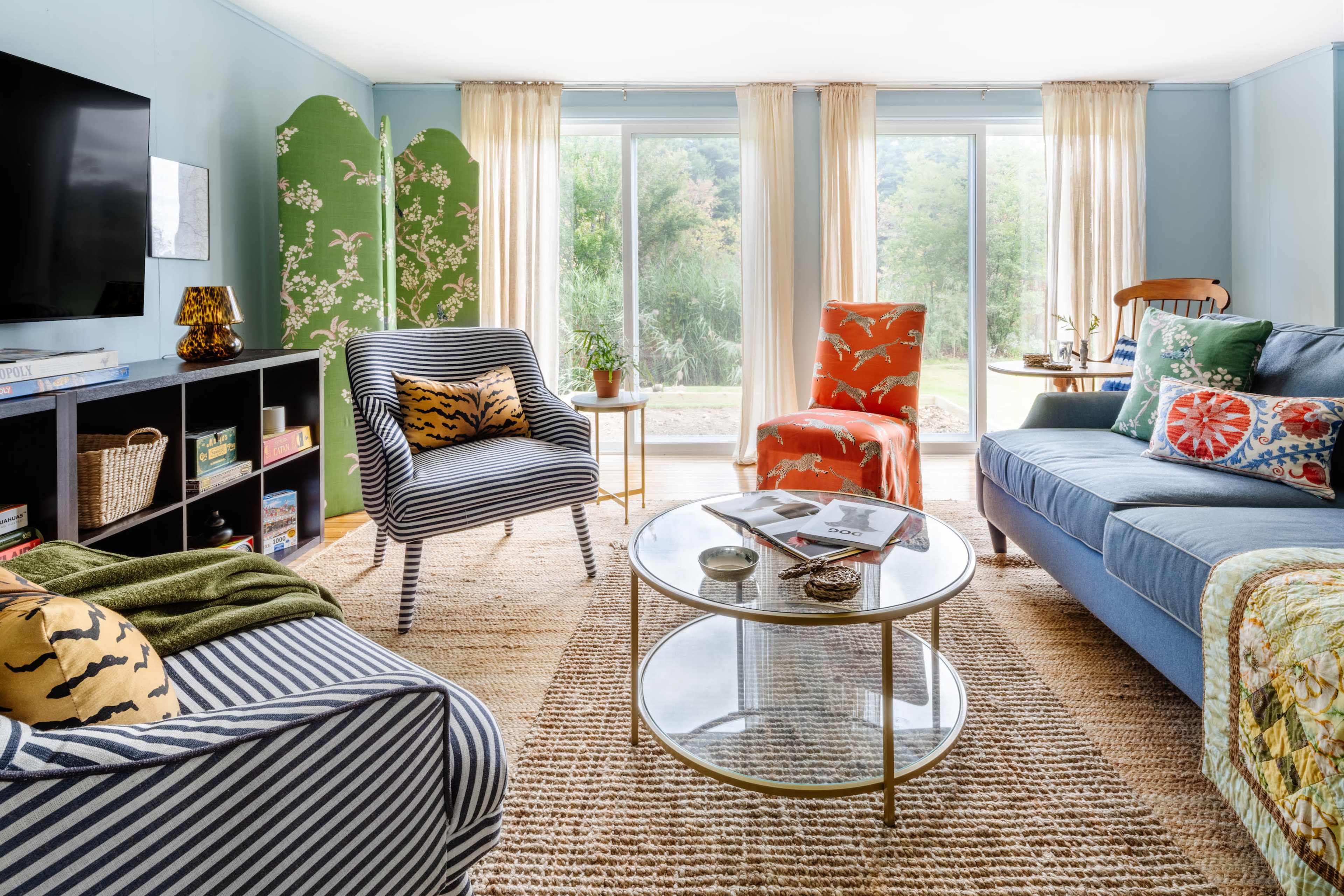 A living room features a blue sofa, patterned armchairs, a round coffee table, and large windows overlooking a garden.