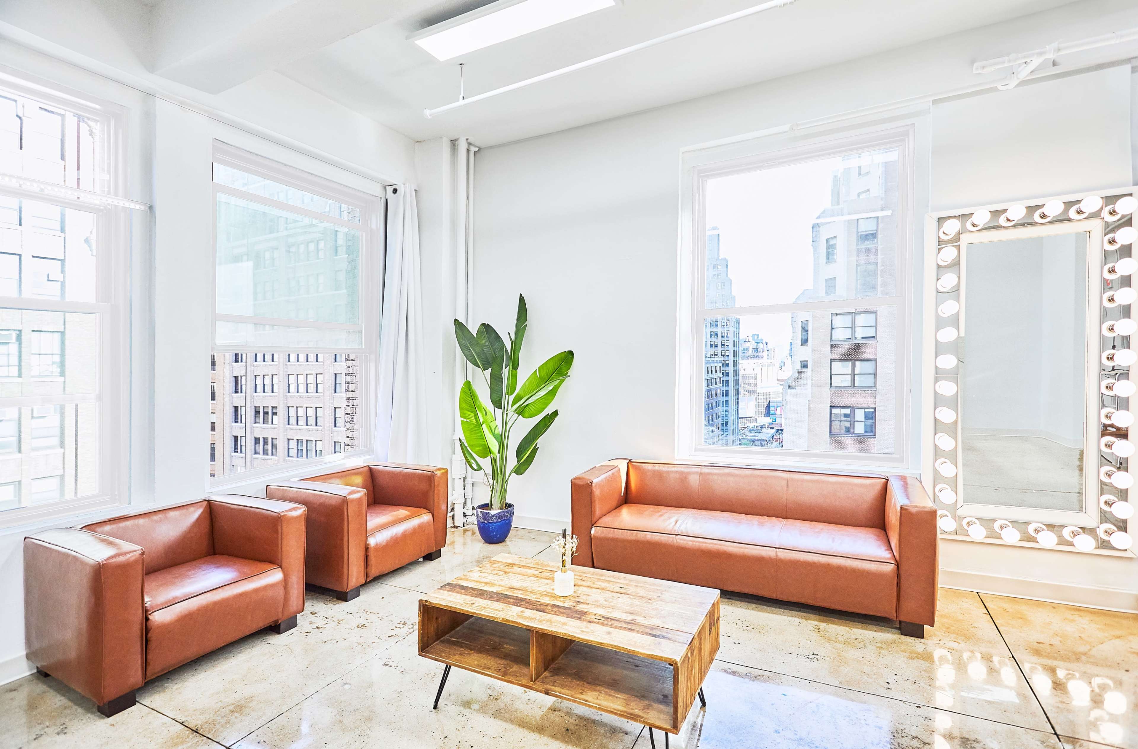 The interior features two brown leather armchairs, a wooden coffee table, and a large mirror with lights, all set against large windows that provide a city view.