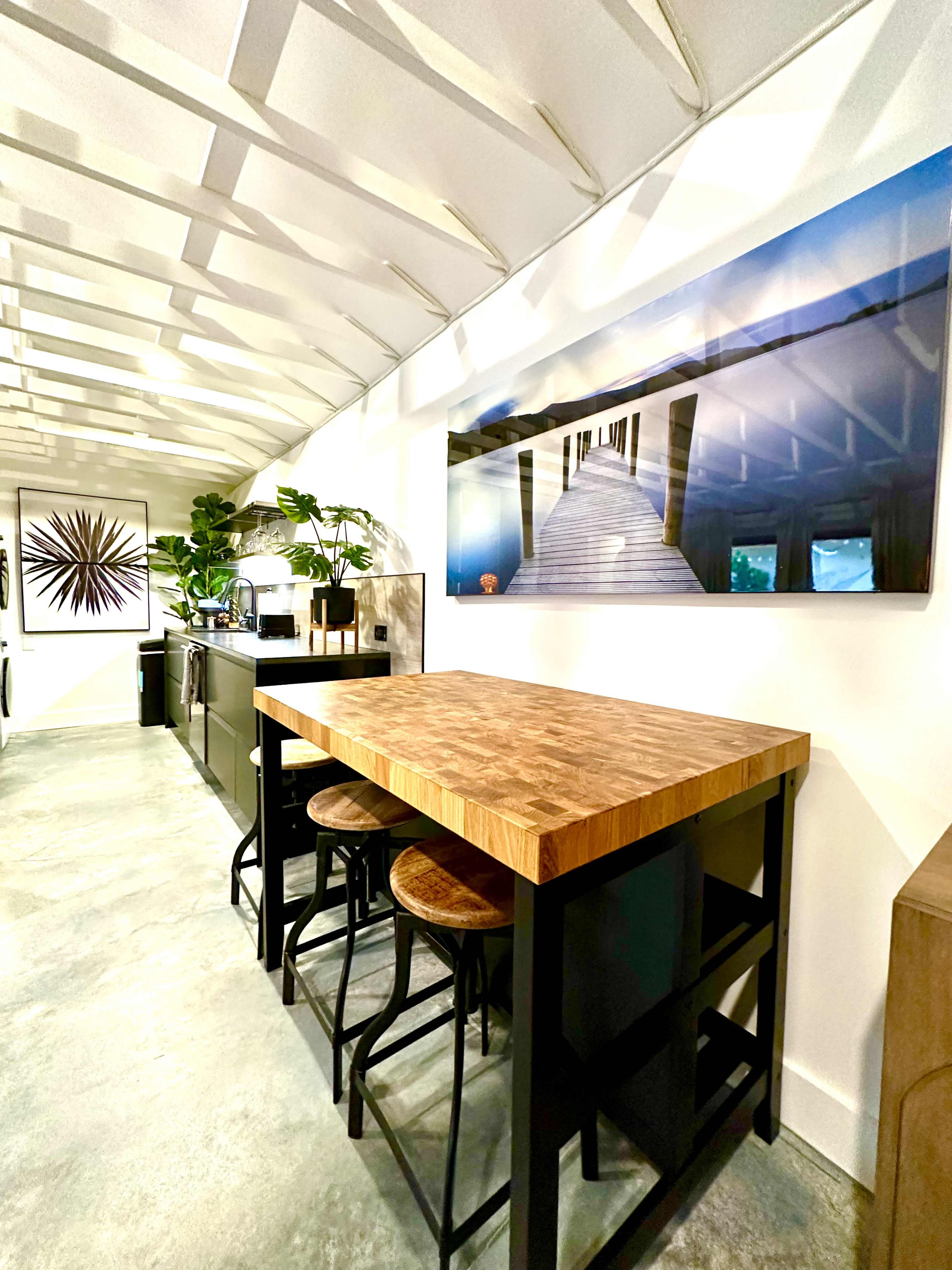 A modern kitchen with a wooden table and stools, framed by white walls and a textured ceiling with plants and artwork on display.