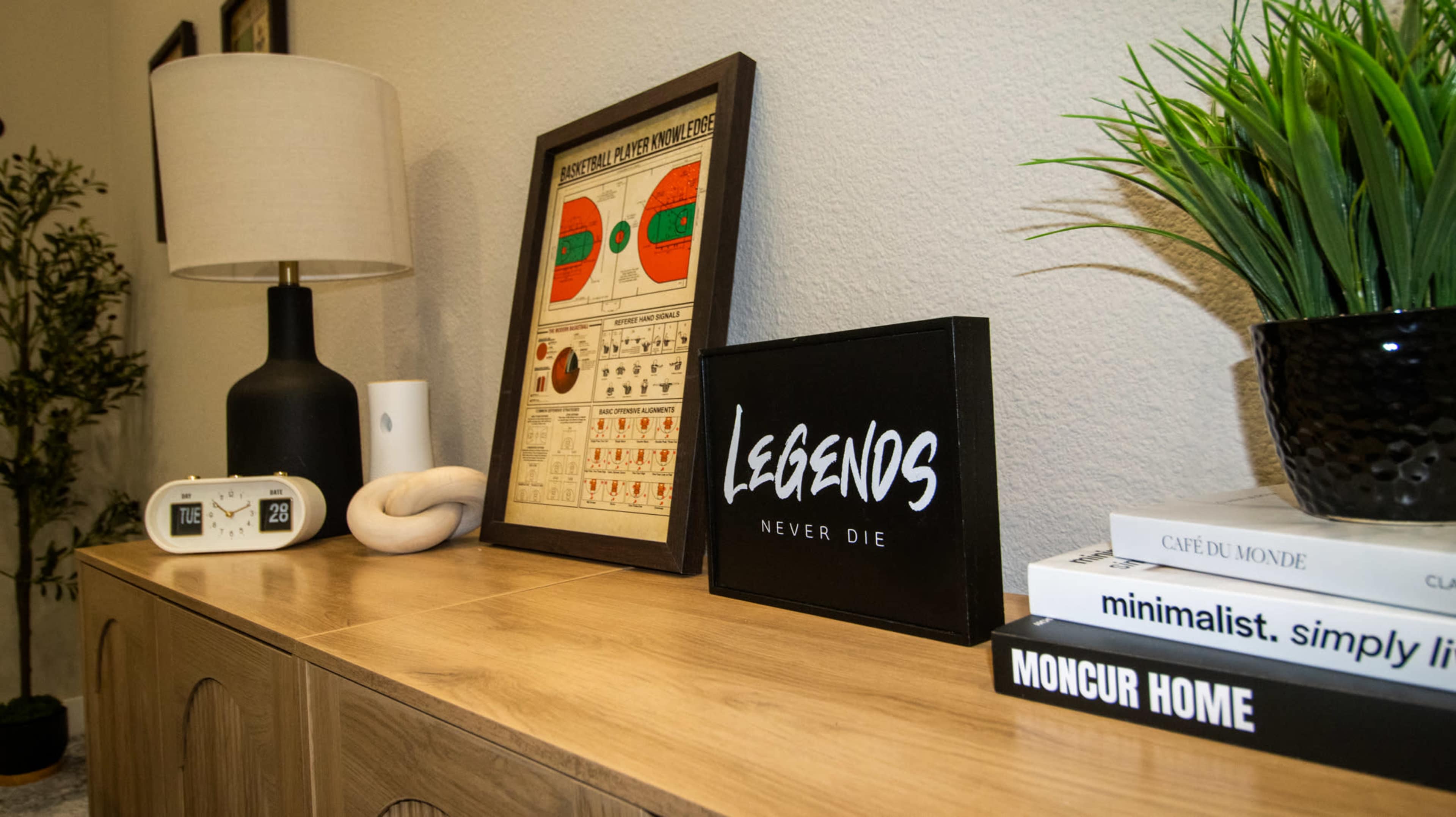 The image shows a wooden sideboard displaying a framed chart, a decorative black sign, a clock, a white sculpture, and stacked books, alongside a green potted plant.
