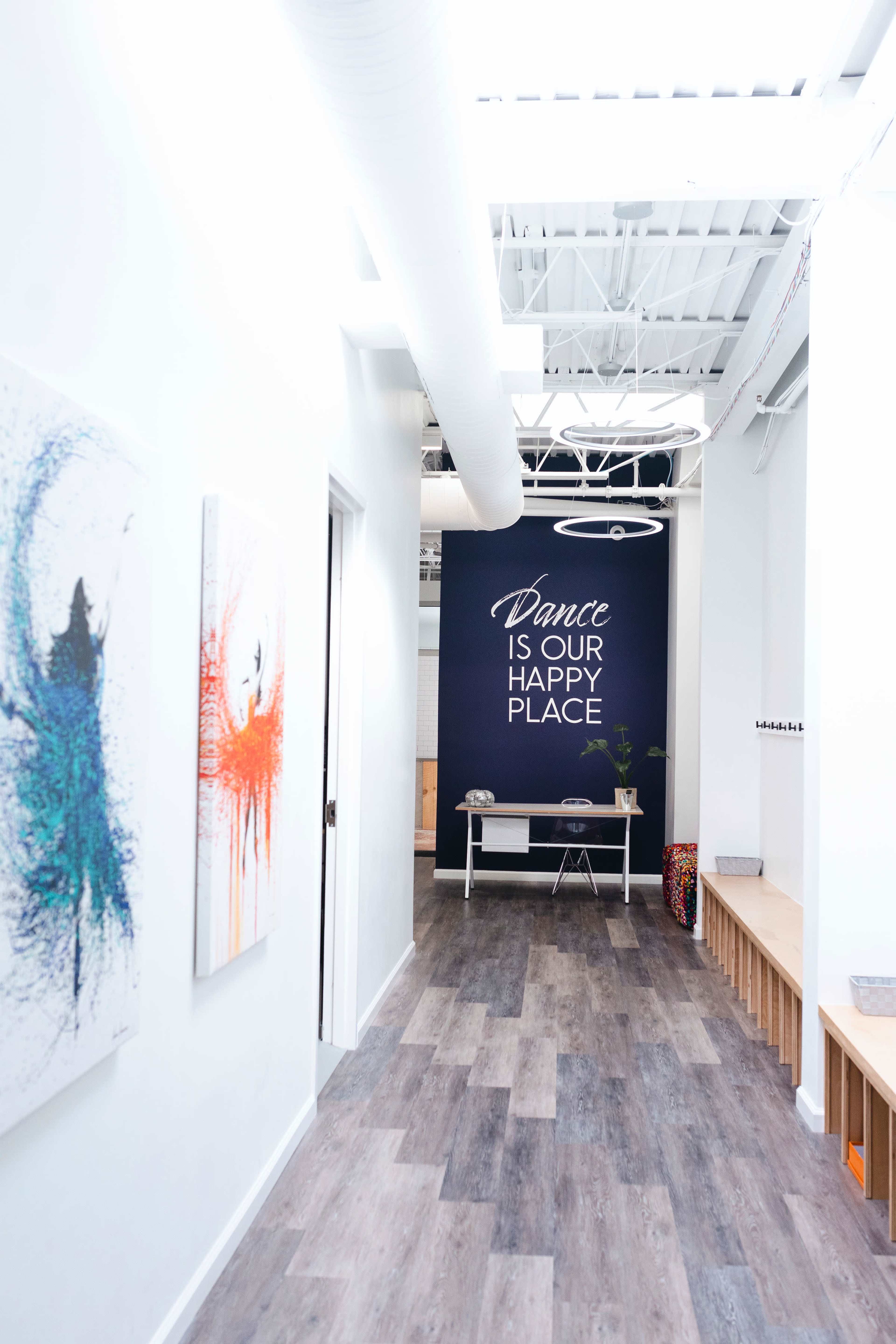 A hallway leading to a room with a wall that has the phrase "Dance Is Our Happy Place" written on it, flanked by colorful artwork and wooden benches along one side.