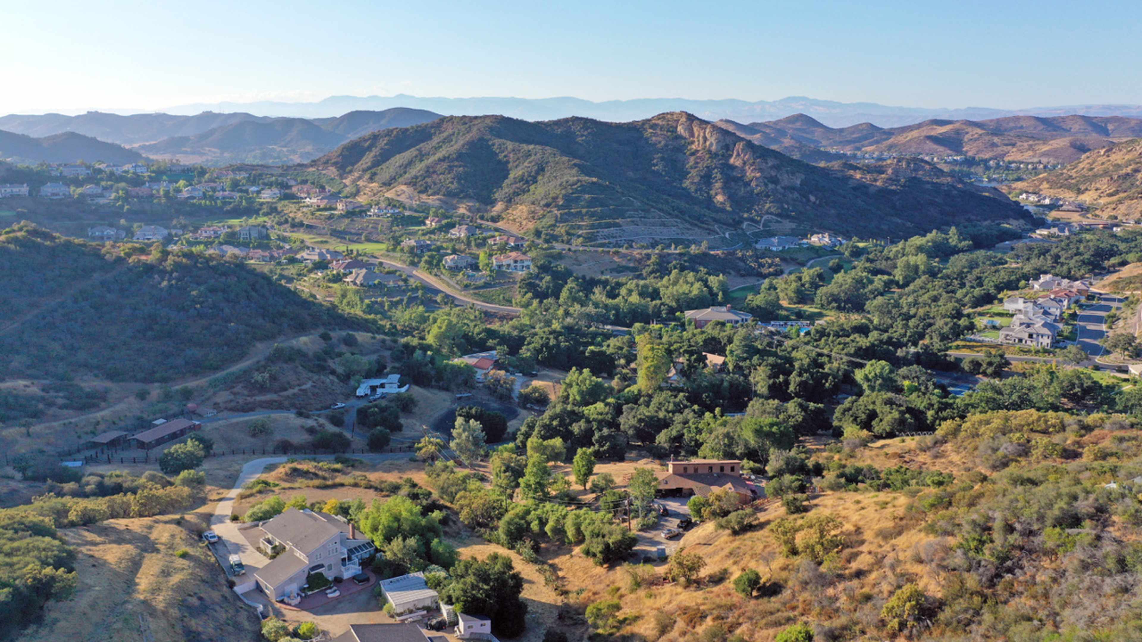 The image shows a wide valley with rolling hills, houses, and patches of greenery under a clear sky.