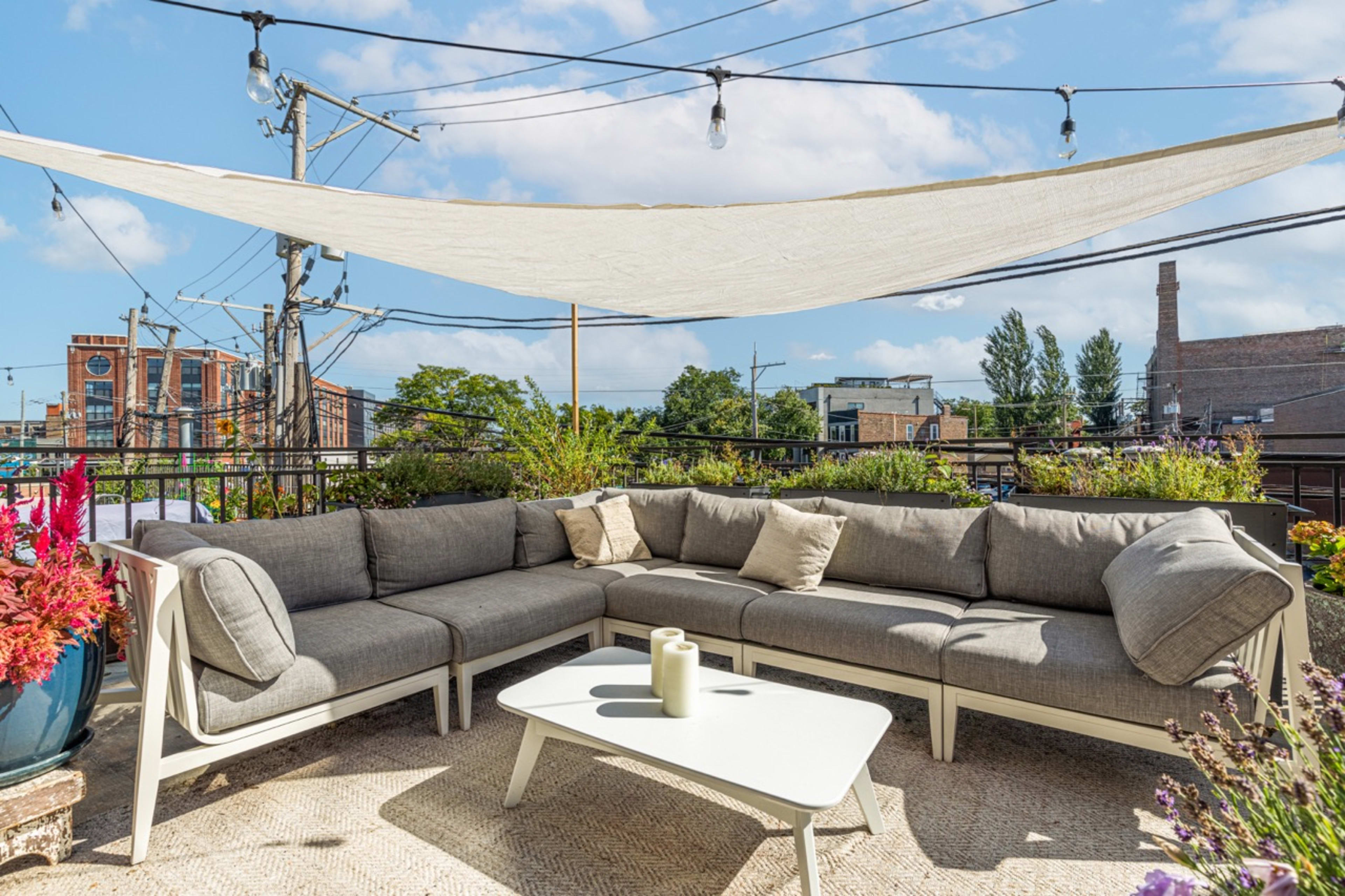 A landscaped rooftop seating area features a modular gray couch, a small table, and a canopy overhead, surrounded by plants and decorative lights.