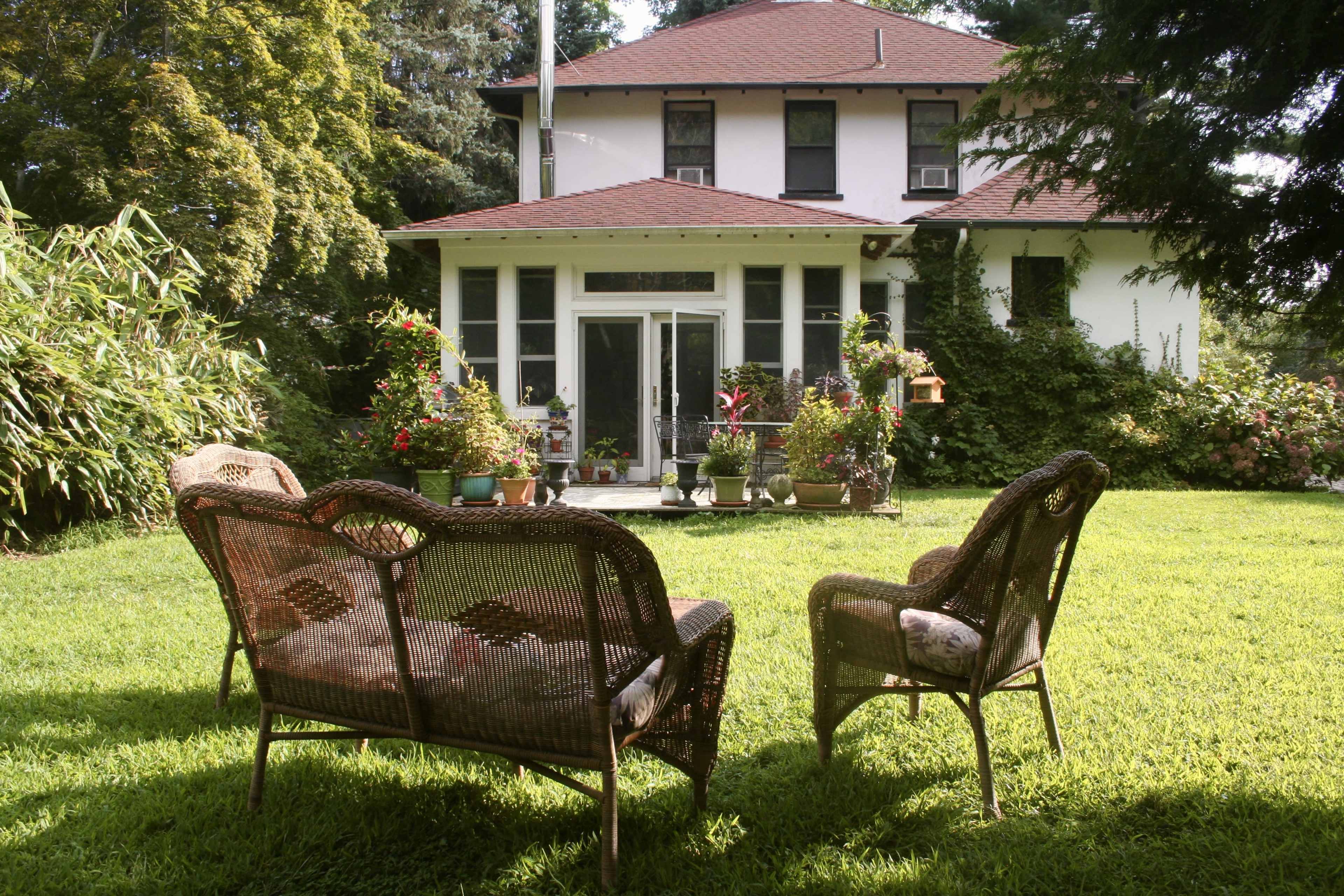 A garden with two wicker chairs in the foreground, facing a white house with a red roof and surrounded by greenery.