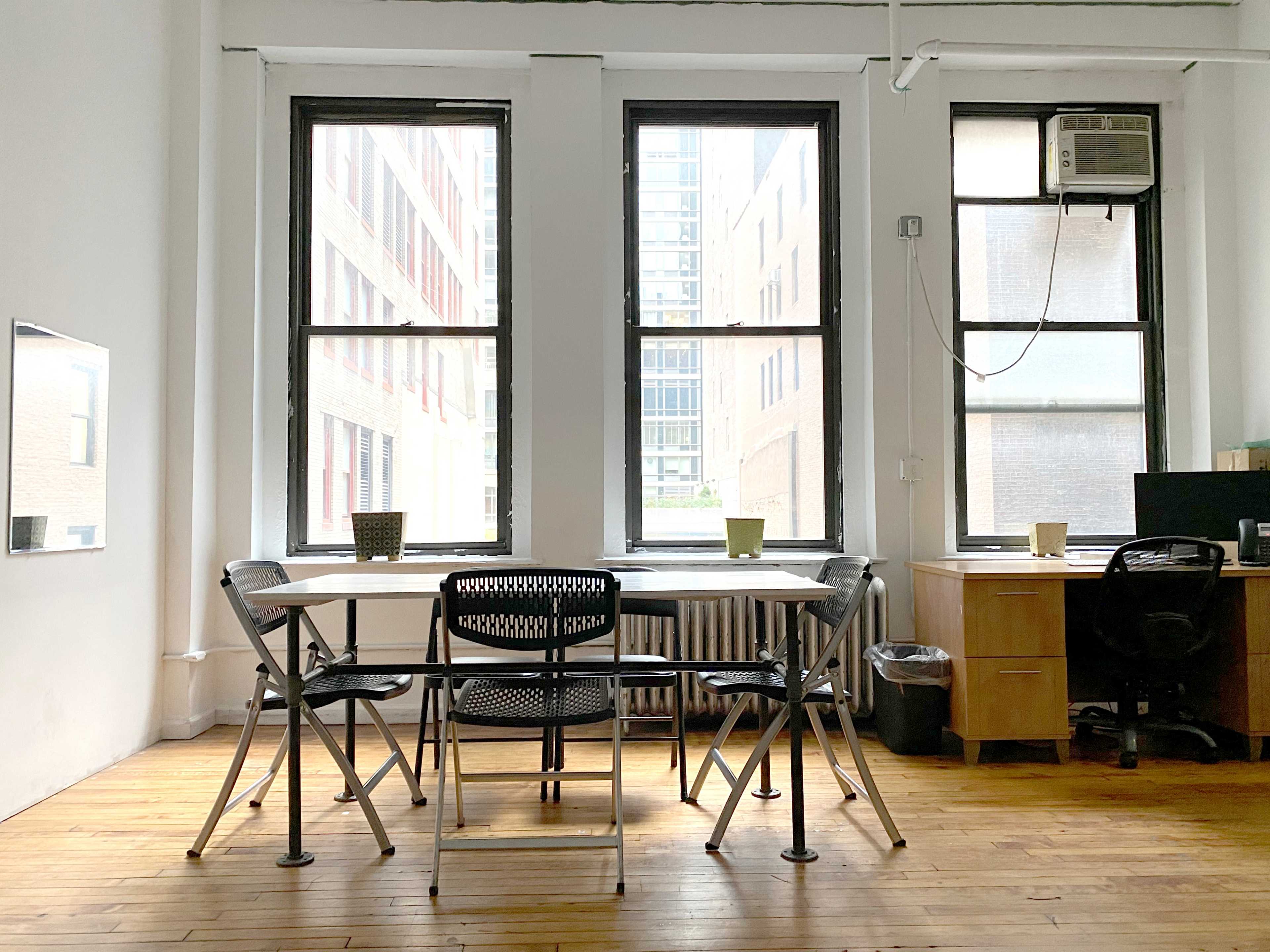 A modern office space features a table with four chairs, two potted plants on the table, and large windows overlooking a cityscape.
