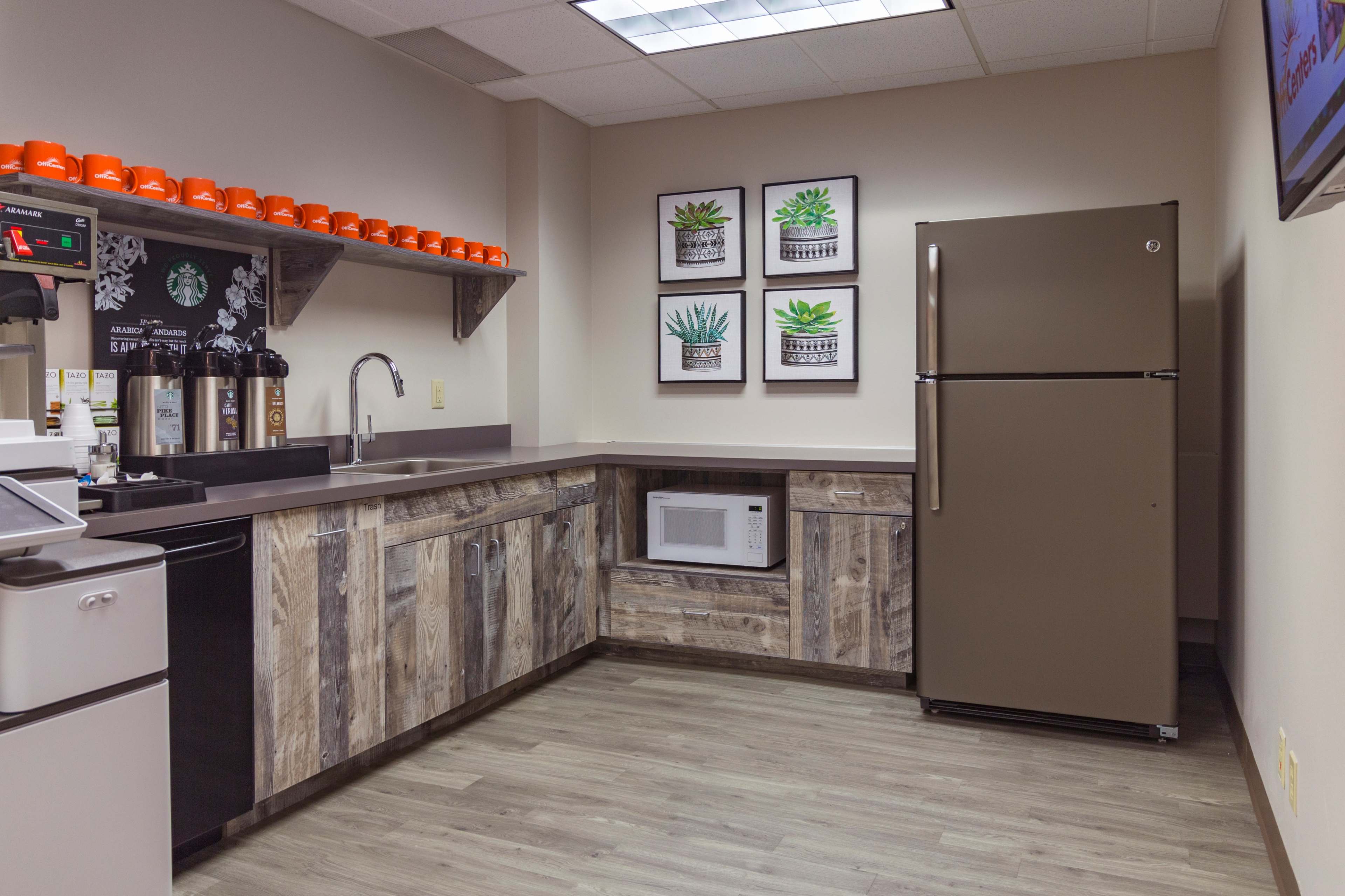A modern break room with a refrigerator, microwave, and coffee station, featuring wooden shelves and framed plant artworks on the wall.