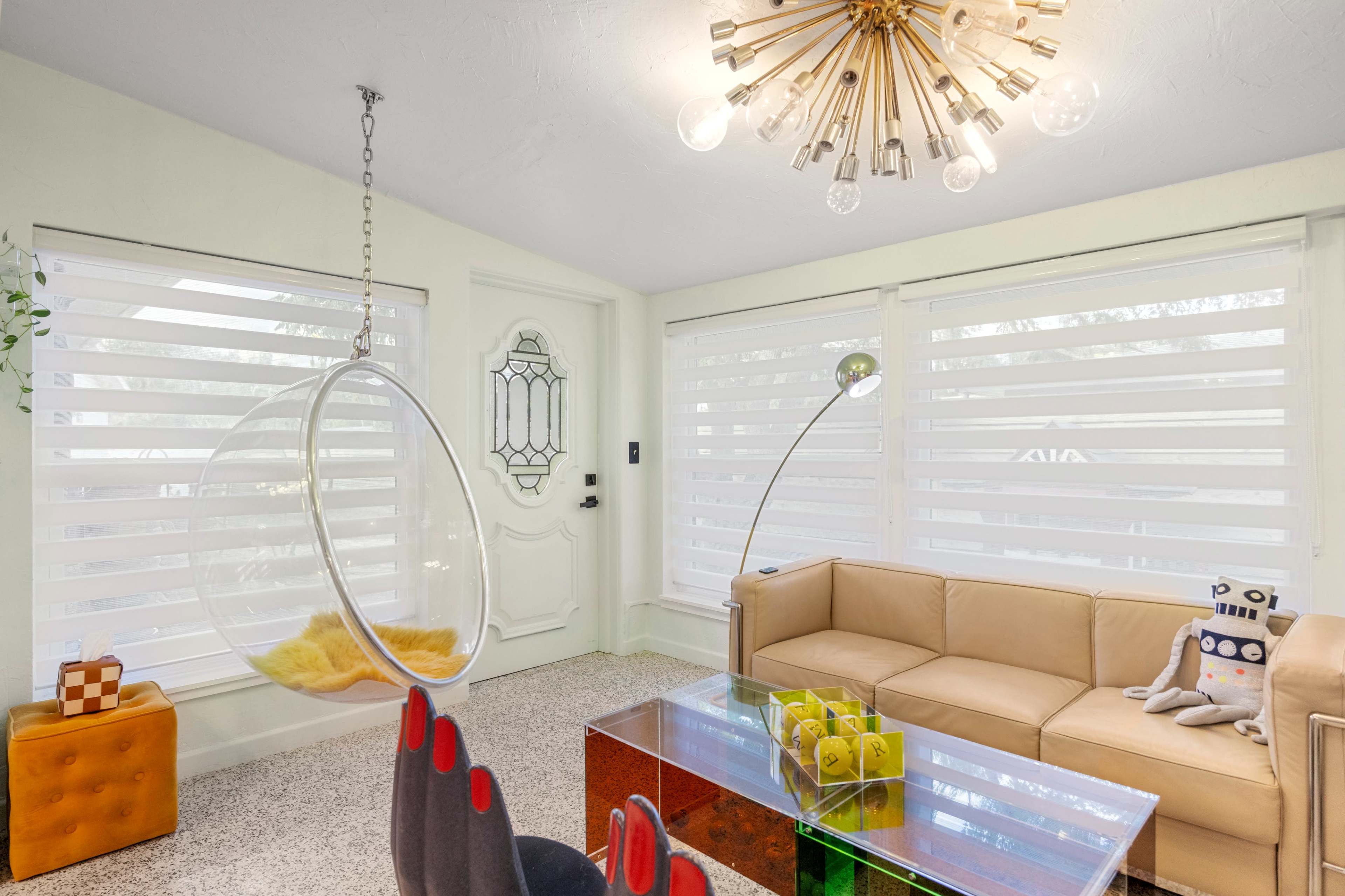 A modern living room with a hanging bubble chair, a tan leather sofa, and a clear glass coffee table, complemented by unique decorative elements and large windows with sheer blinds.