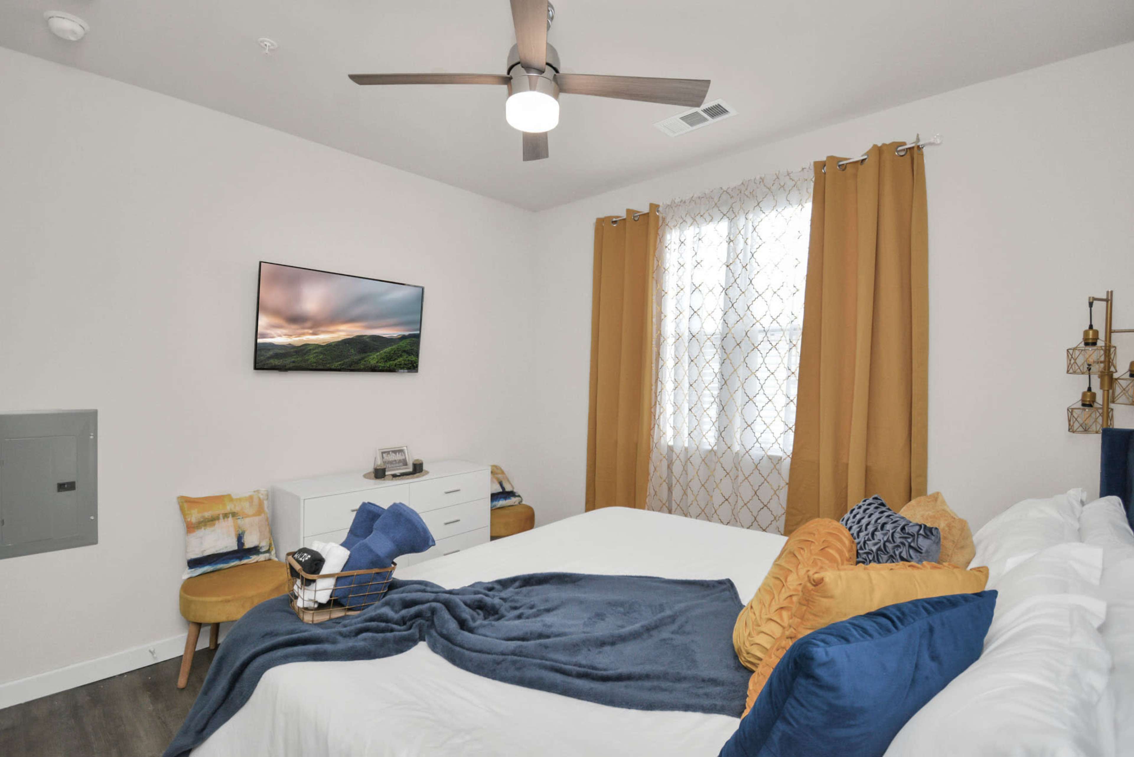 The image shows a neatly arranged bedroom featuring a white bed, a television mounted on the wall, and large windows dressed with yellow curtains.