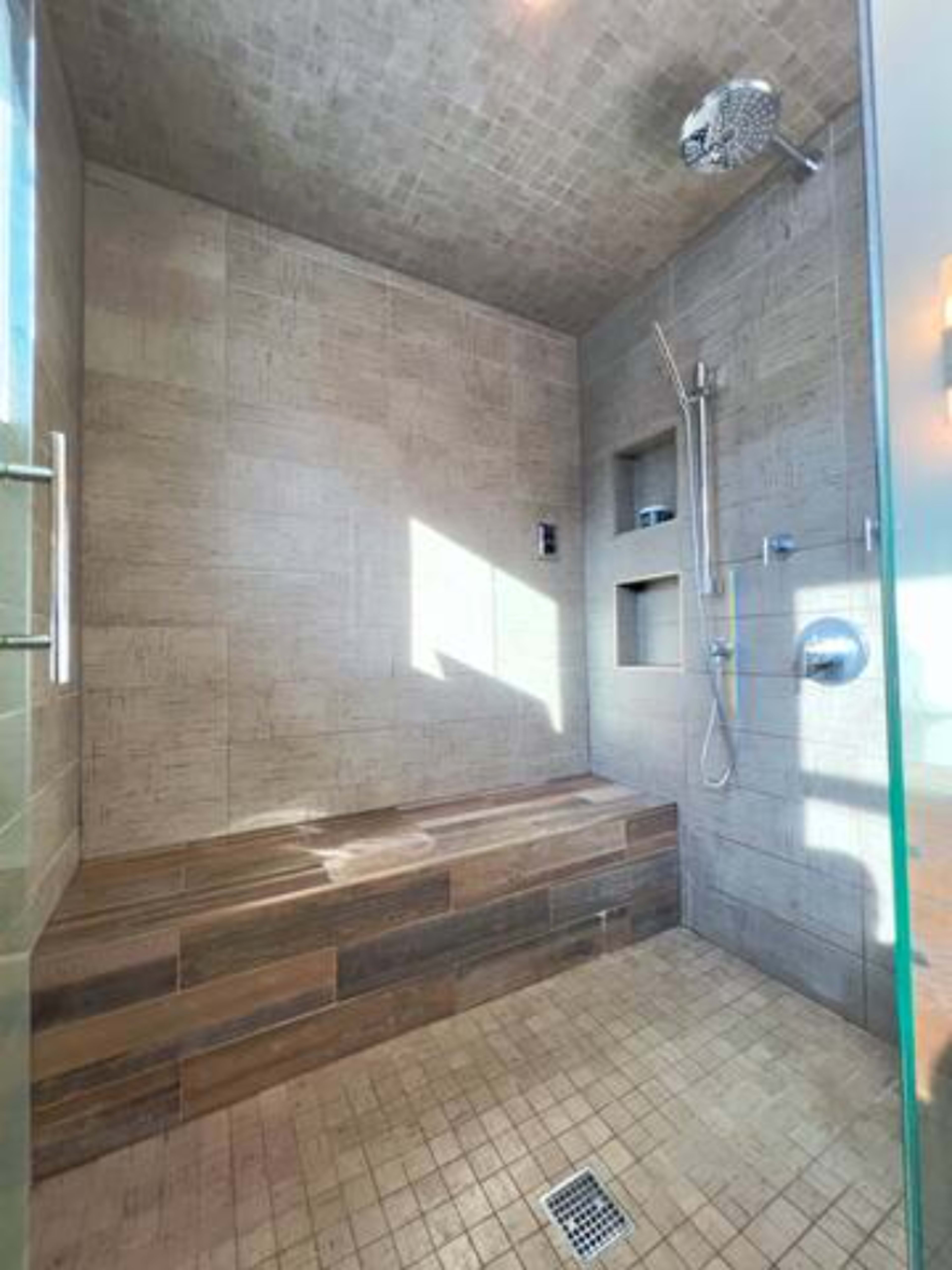 The image shows a modern shower area with tiled walls, a wooden bench, and a large showerhead.