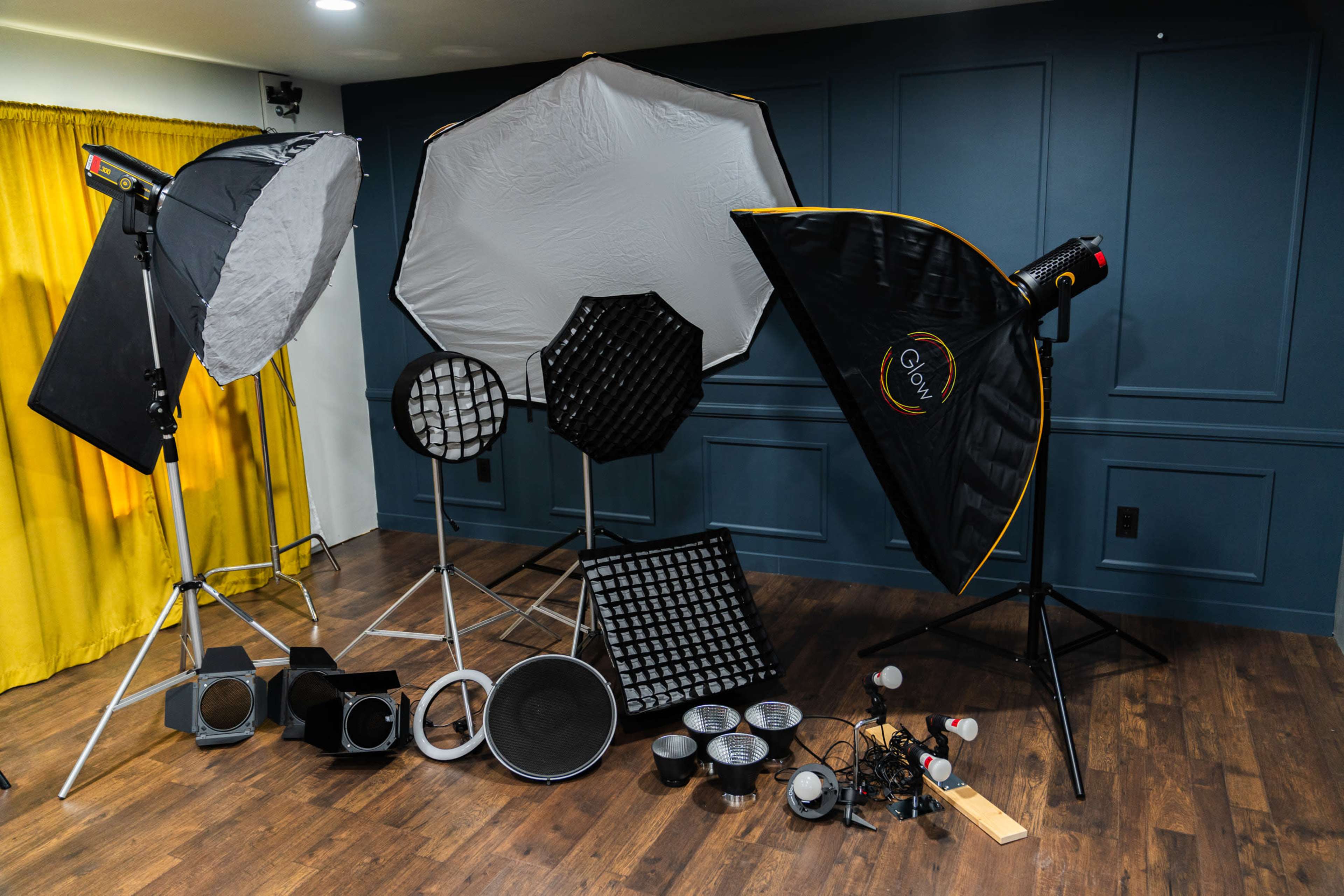 The image shows an assortment of photography lighting equipment arranged against a dark blue wall, including softboxes, reflectors, and various light modifiers.