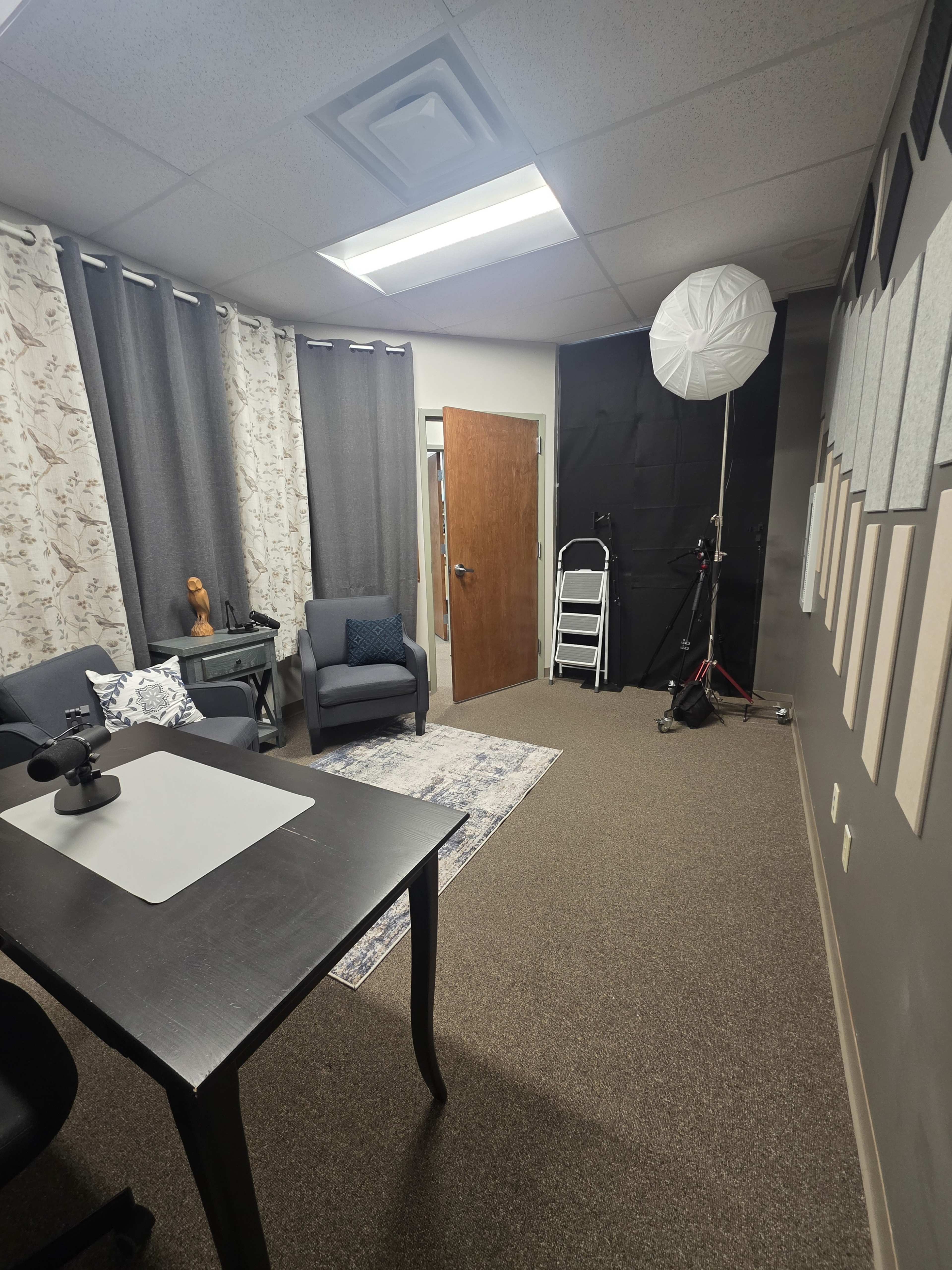 The image shows a small office space featuring a desk with a chair, two armchairs, curtains, and a backdrop setup for photography.