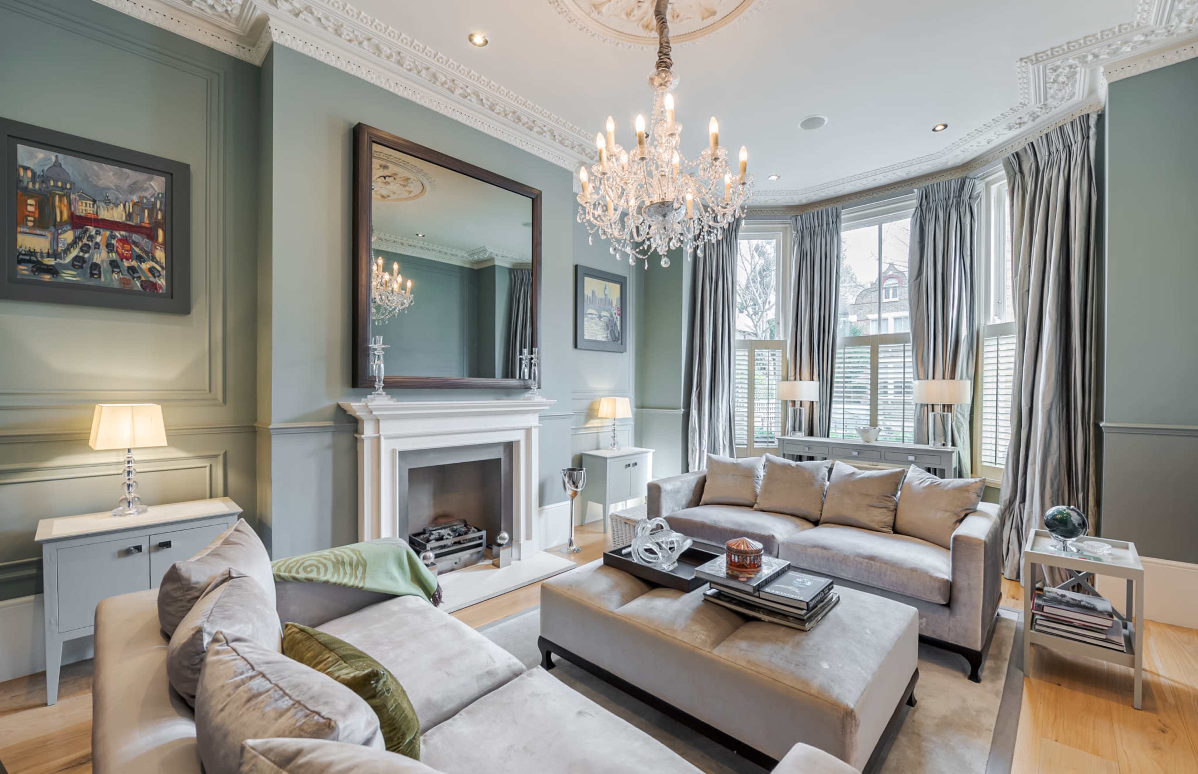 The image shows a stylish living room with elegant decor, featuring a chandelier, a large mirror, a fireplace, gray sofas, and large windows with drapes.