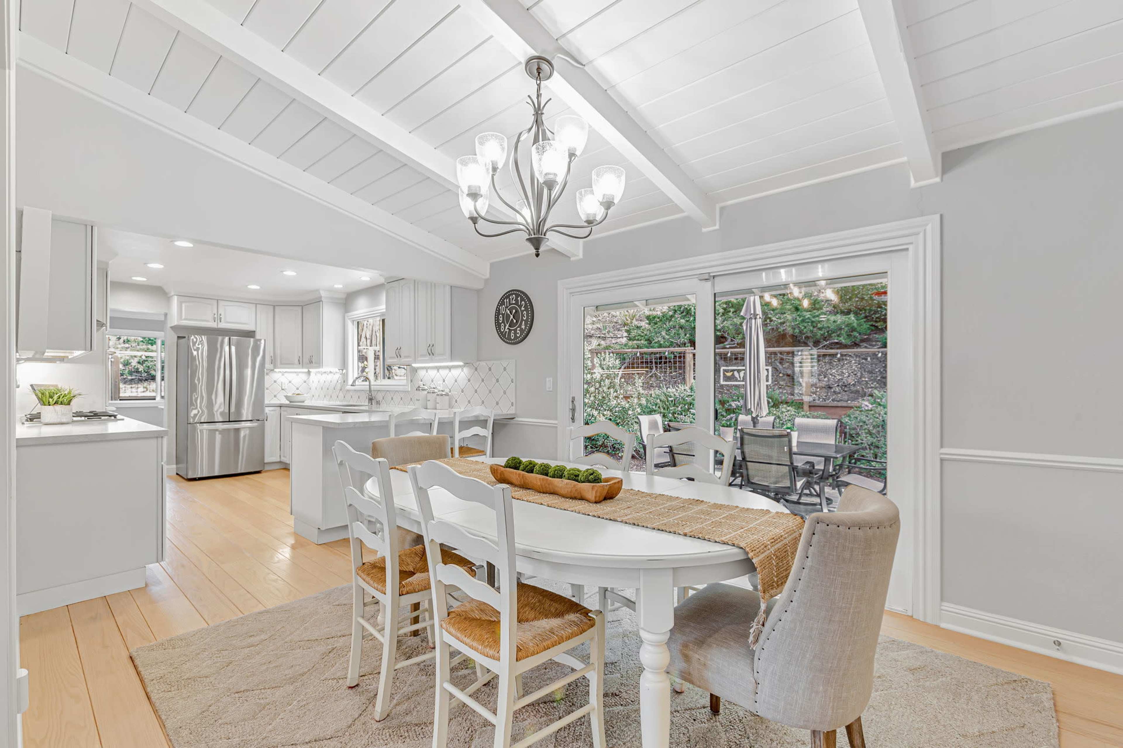 The image depicts a bright, modern dining area with a white table, four chairs, and a chandelier, which opens to an outdoor patio through sliding glass doors.