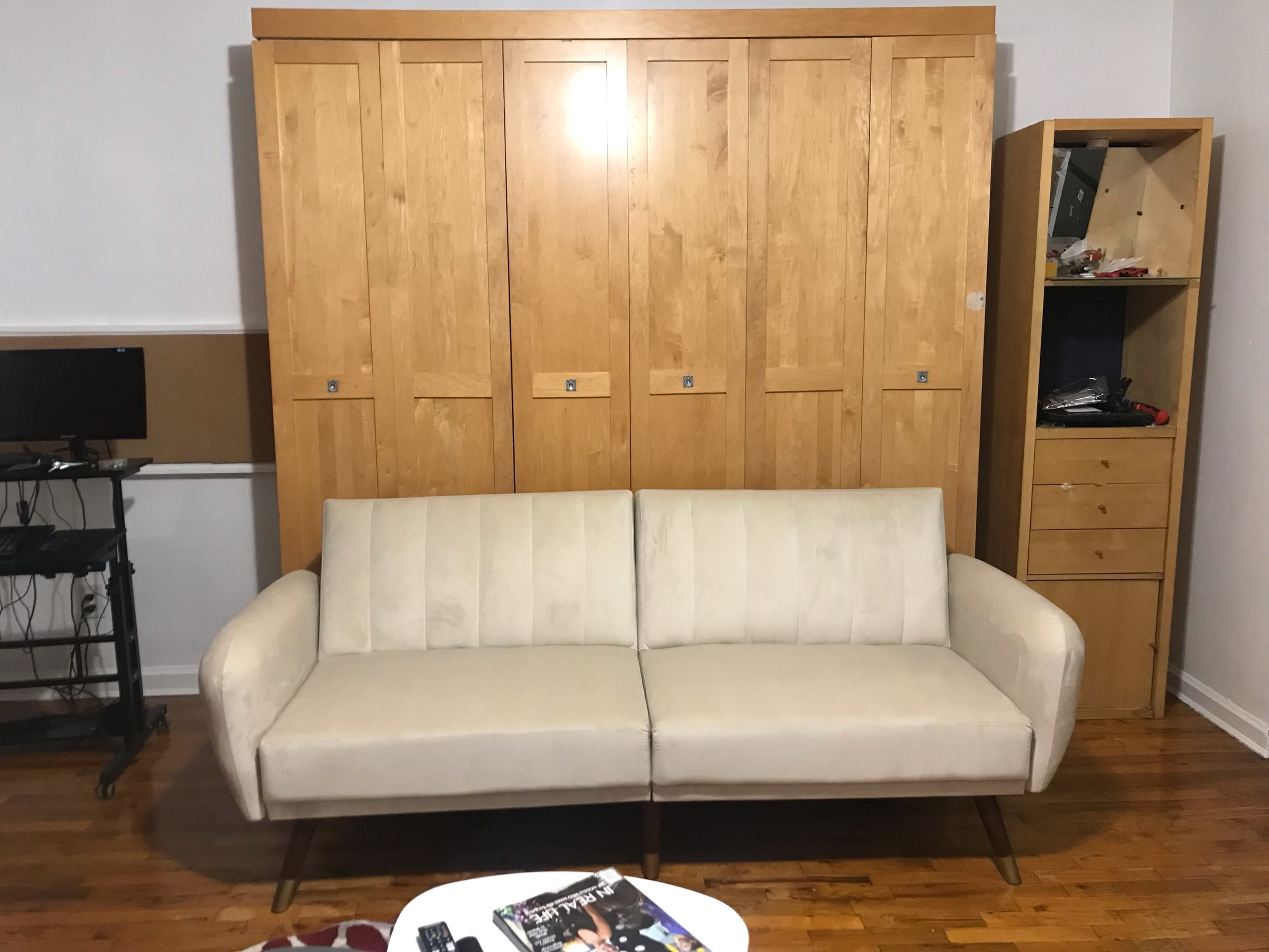 The image shows a light-colored sofa positioned in front of a wooden wall unit with multiple doors and a small shelving unit on the side.