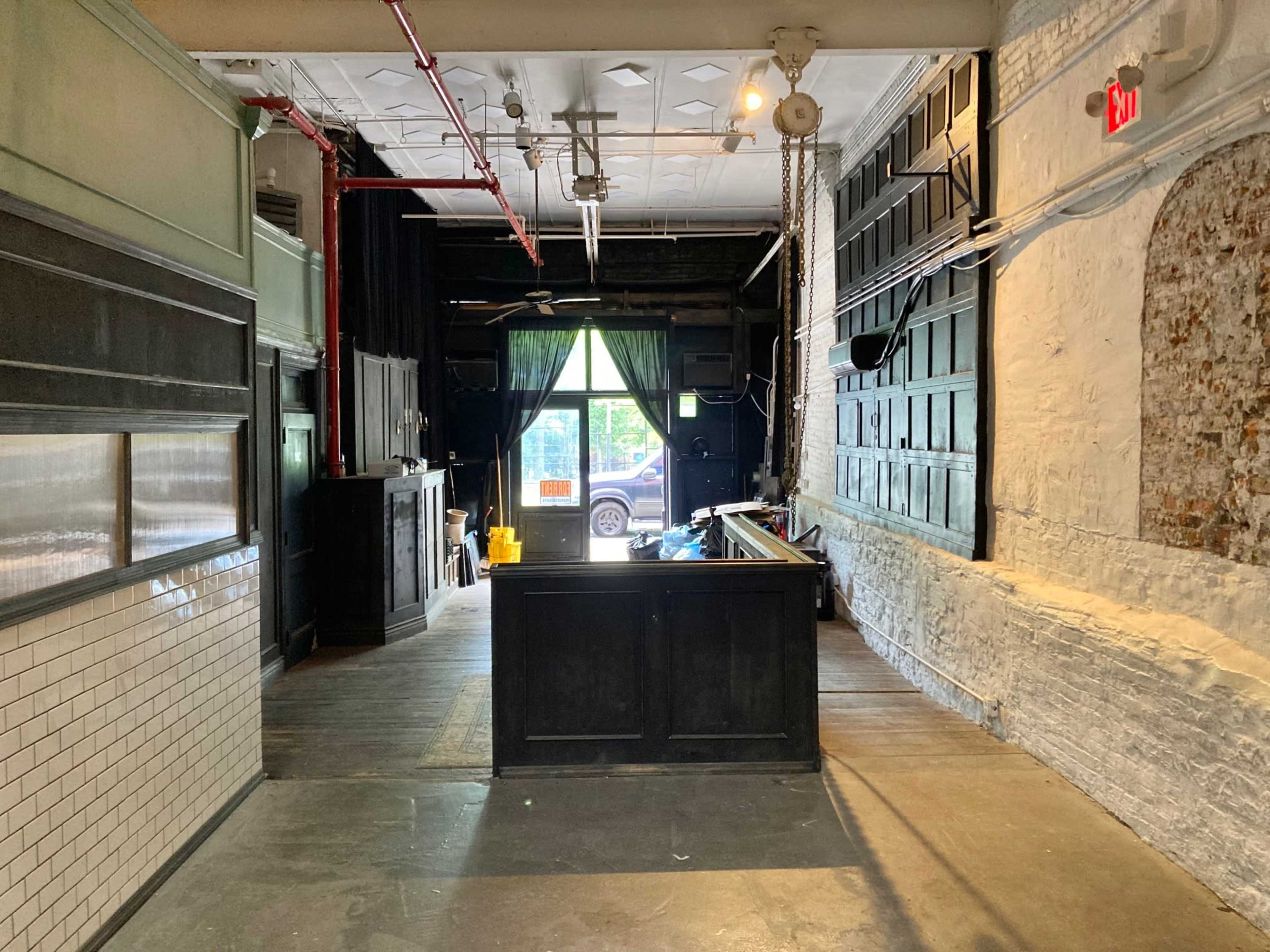 The image shows the interior of a vacant establishment with a bar area near the entrance, exposed brick walls, and green curtains visible through the front window.