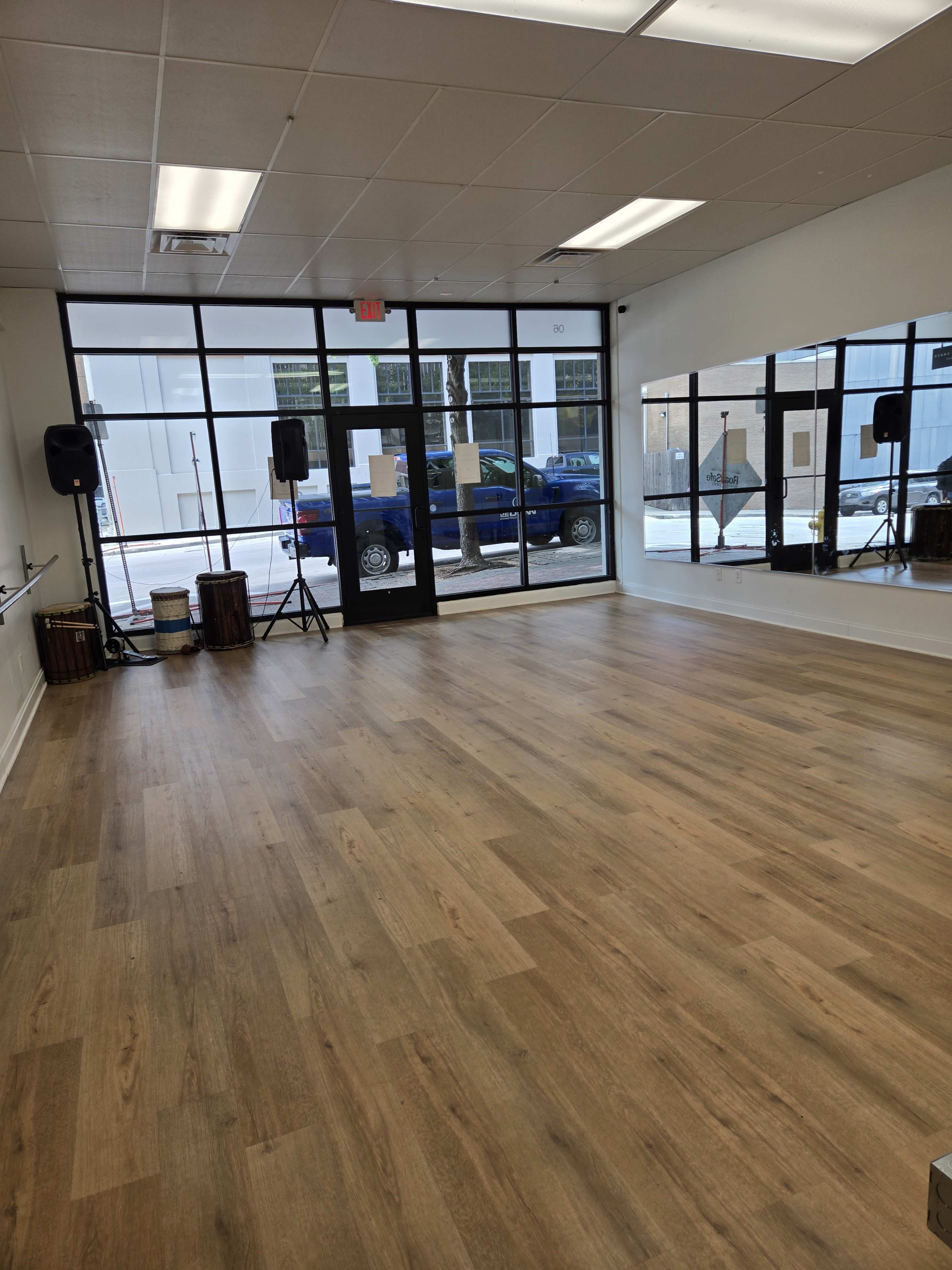 The image shows a spacious, empty dance studio with a wooden floor, large windows, and mirrors along one wall.