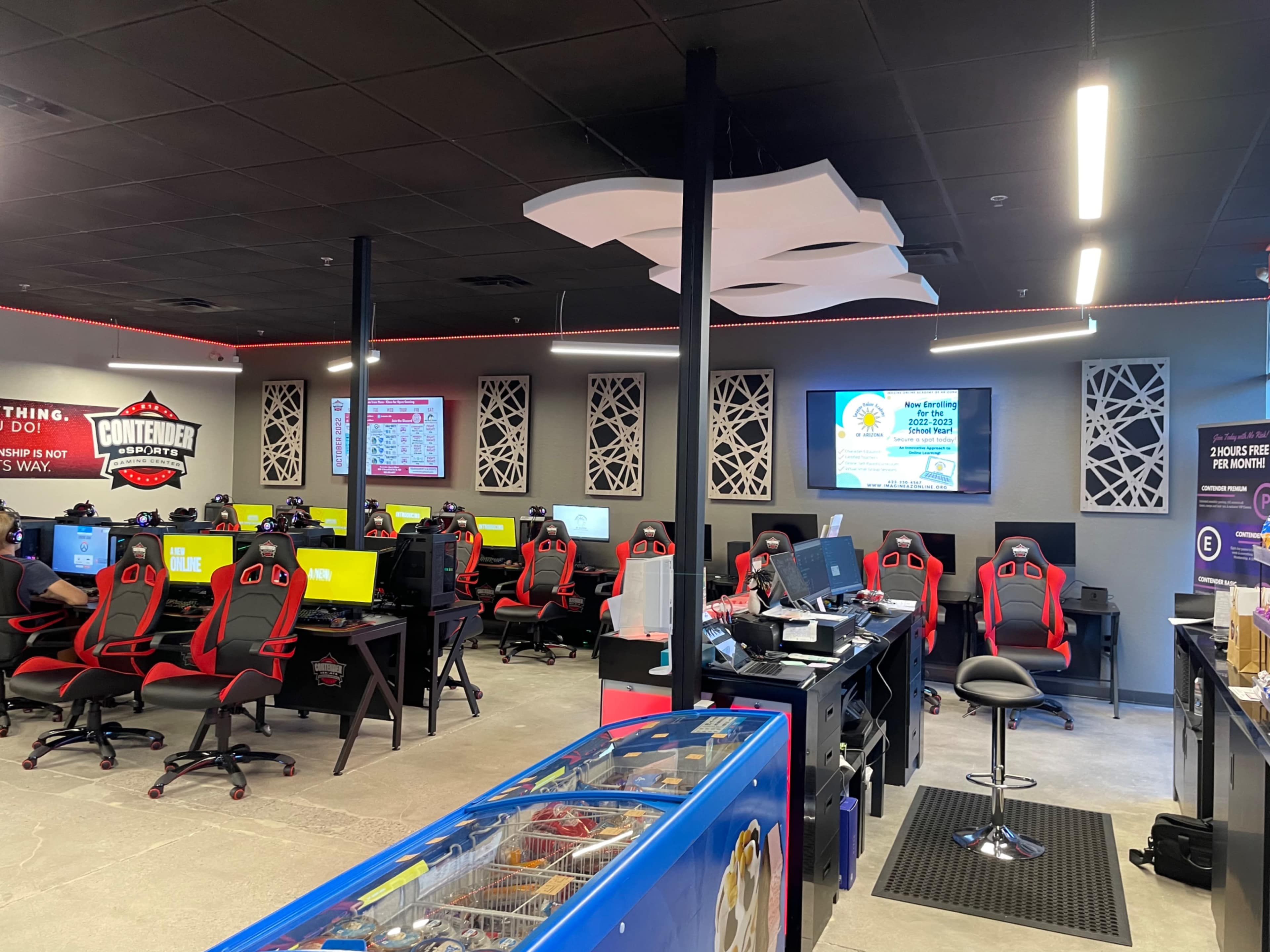 A gaming center with rows of computer workstations featuring red gaming chairs, screens displaying games, and a section with a snack bar.
