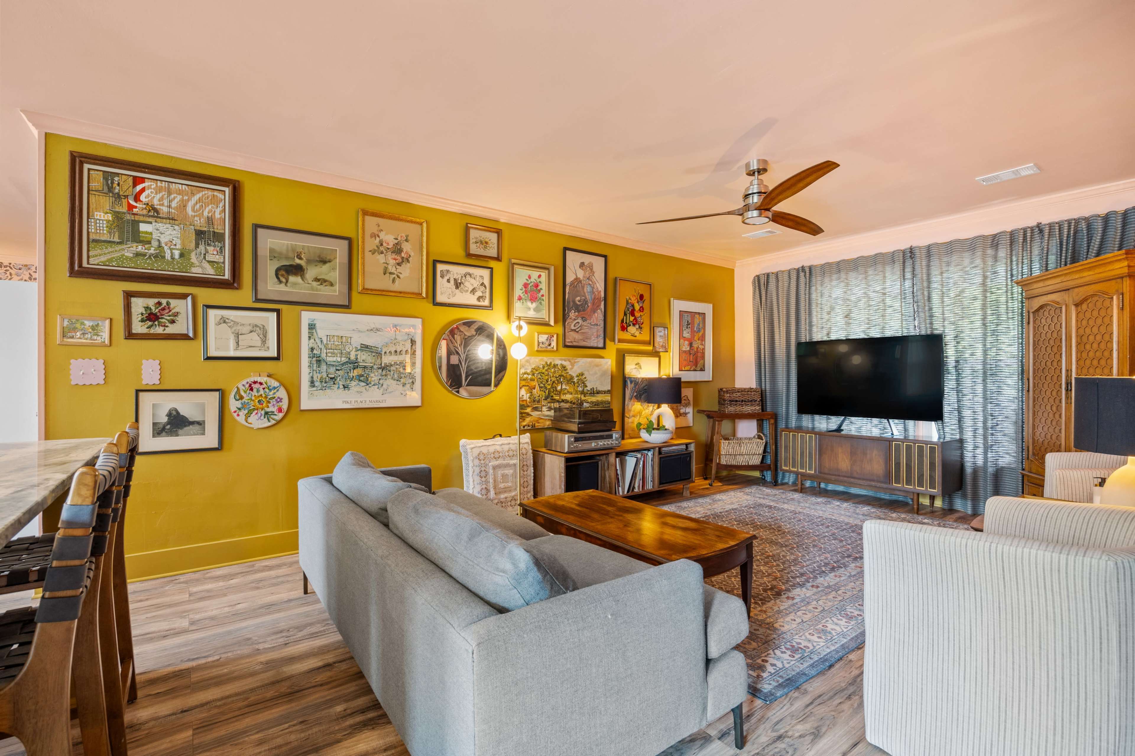 The image shows a cozy living room featuring a wall adorned with various framed artworks, a television, and comfortable seating arrangements.