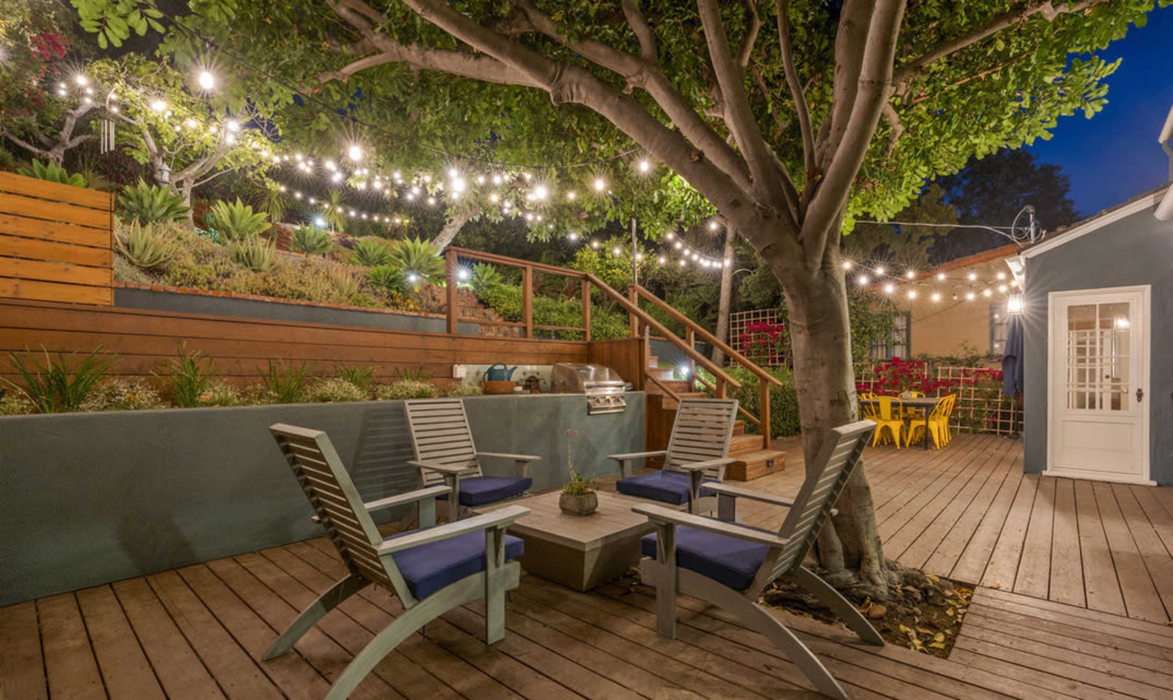A cozy outdoor patio area features wooden chairs arranged around a table under string lights, with a staircase leading to a landscaped garden.