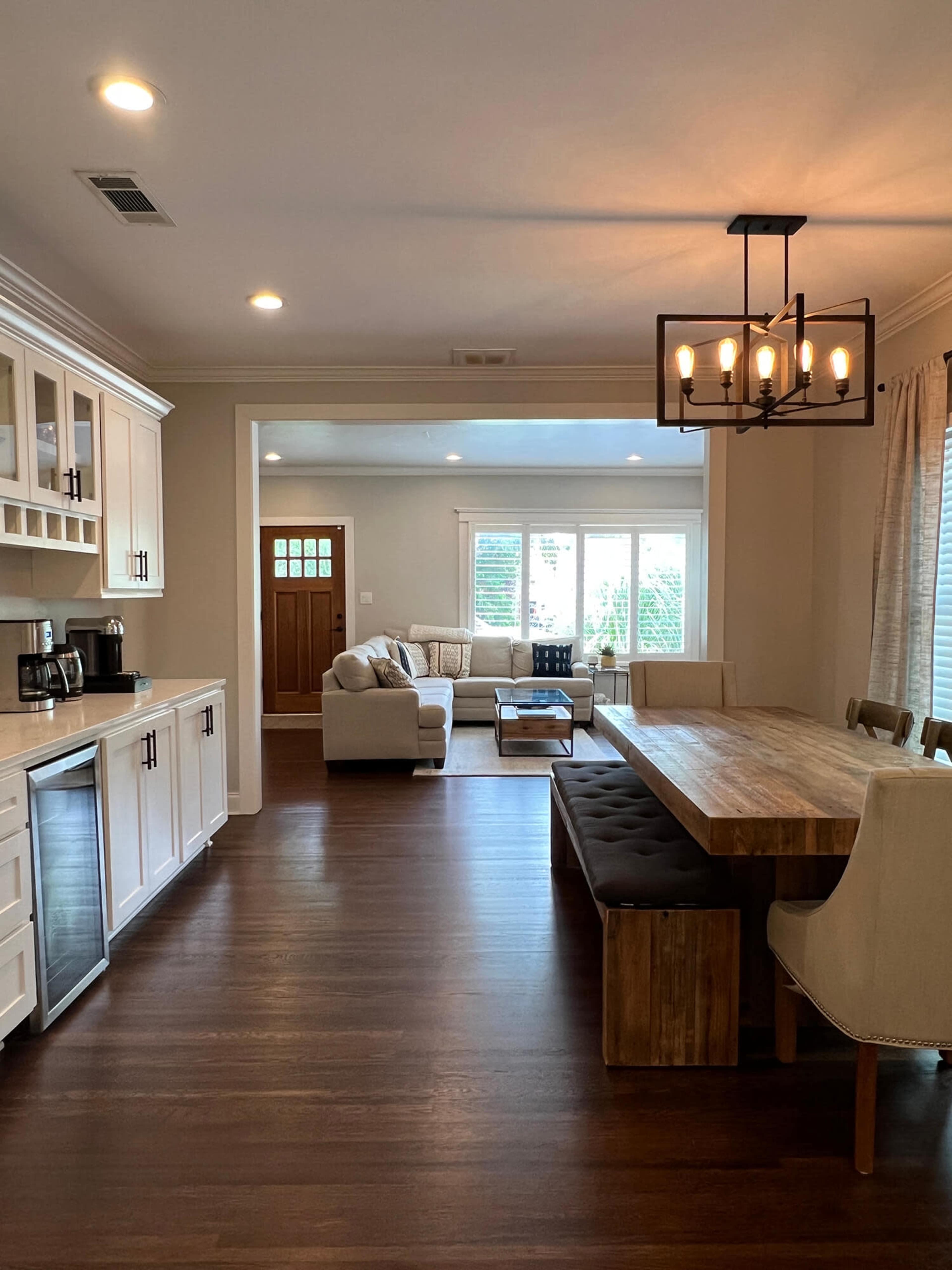 A modern kitchen and dining area features light wood cabinetry, a large wooden dining table, and a comfortable seating area with a sofa and windows providing natural light.