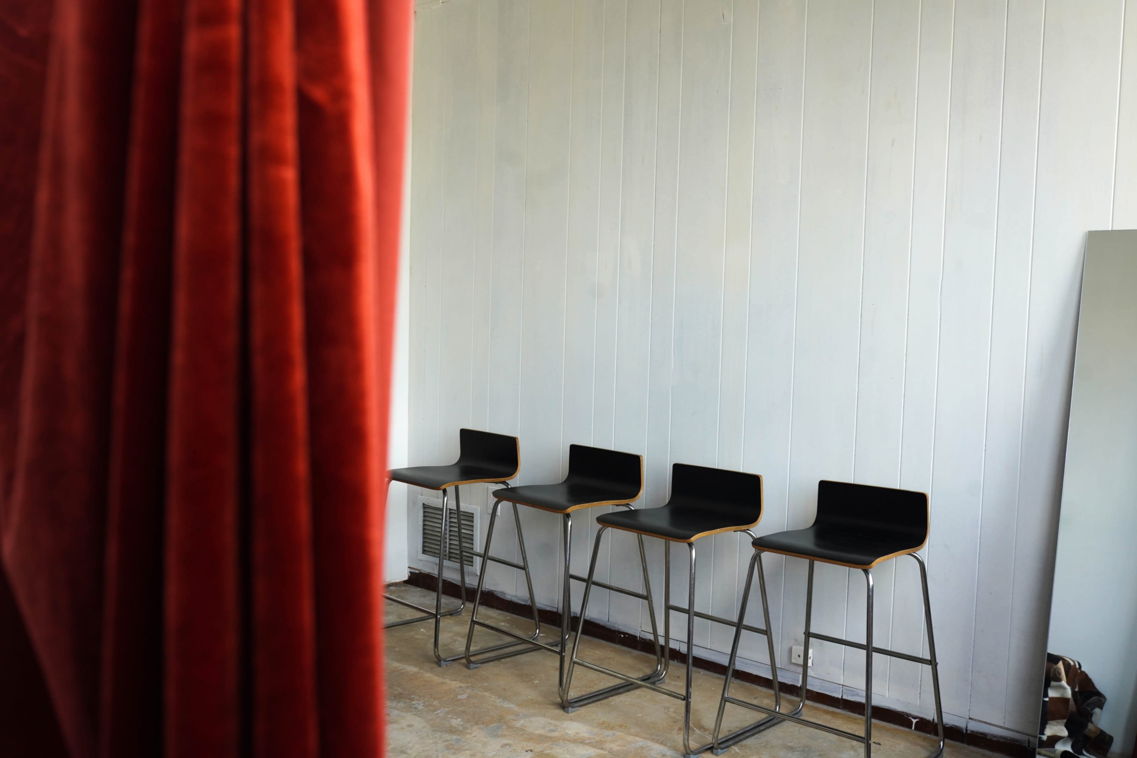 The image shows a sparsely furnished room with four black bar stools arranged against a light-colored wall, partially obscured by a red curtain.