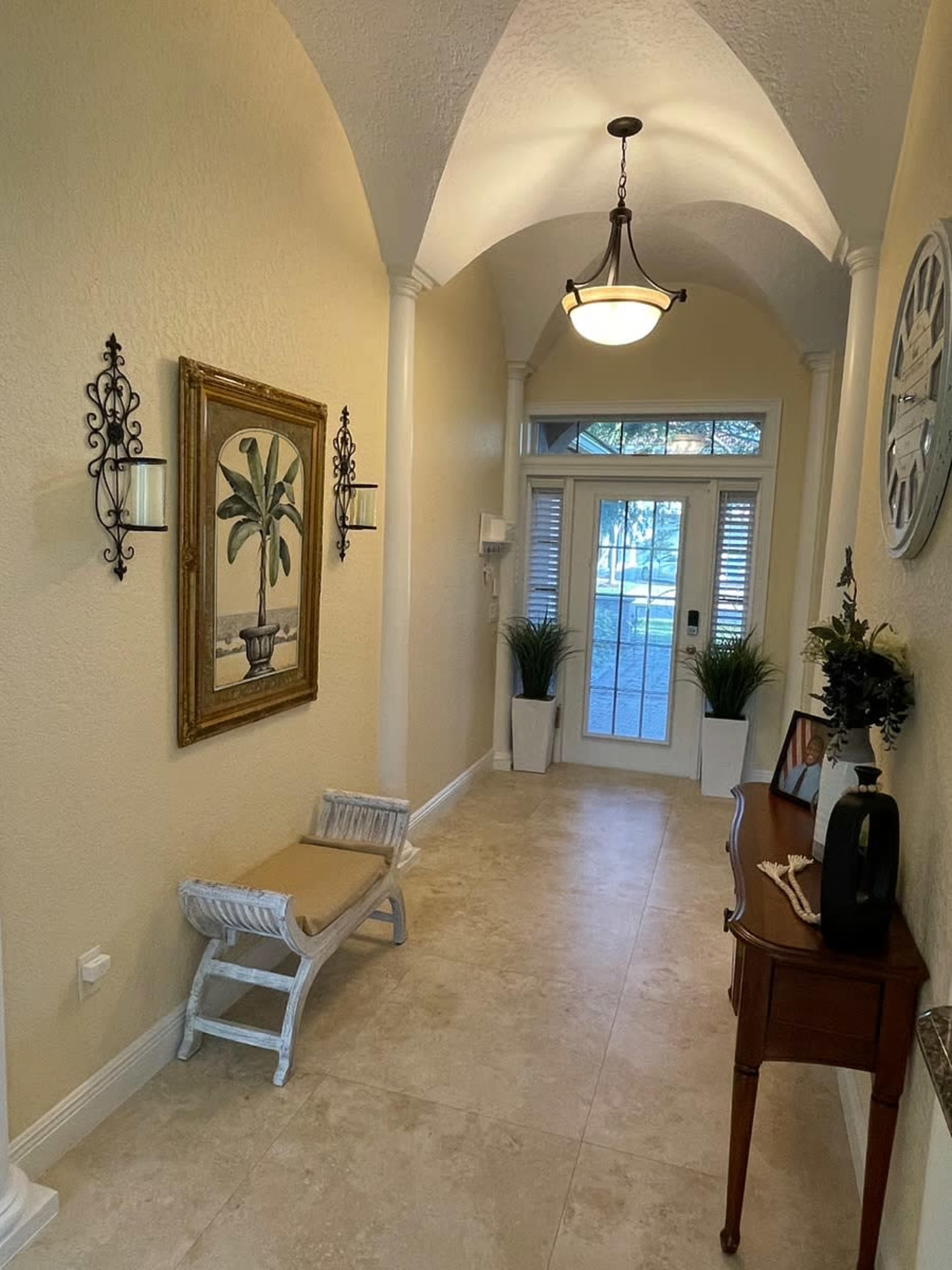The image shows a spacious hallway with a decorative painting, wall sconces, potted plants, and a small wooden table, leading to a glass-paneled front door.