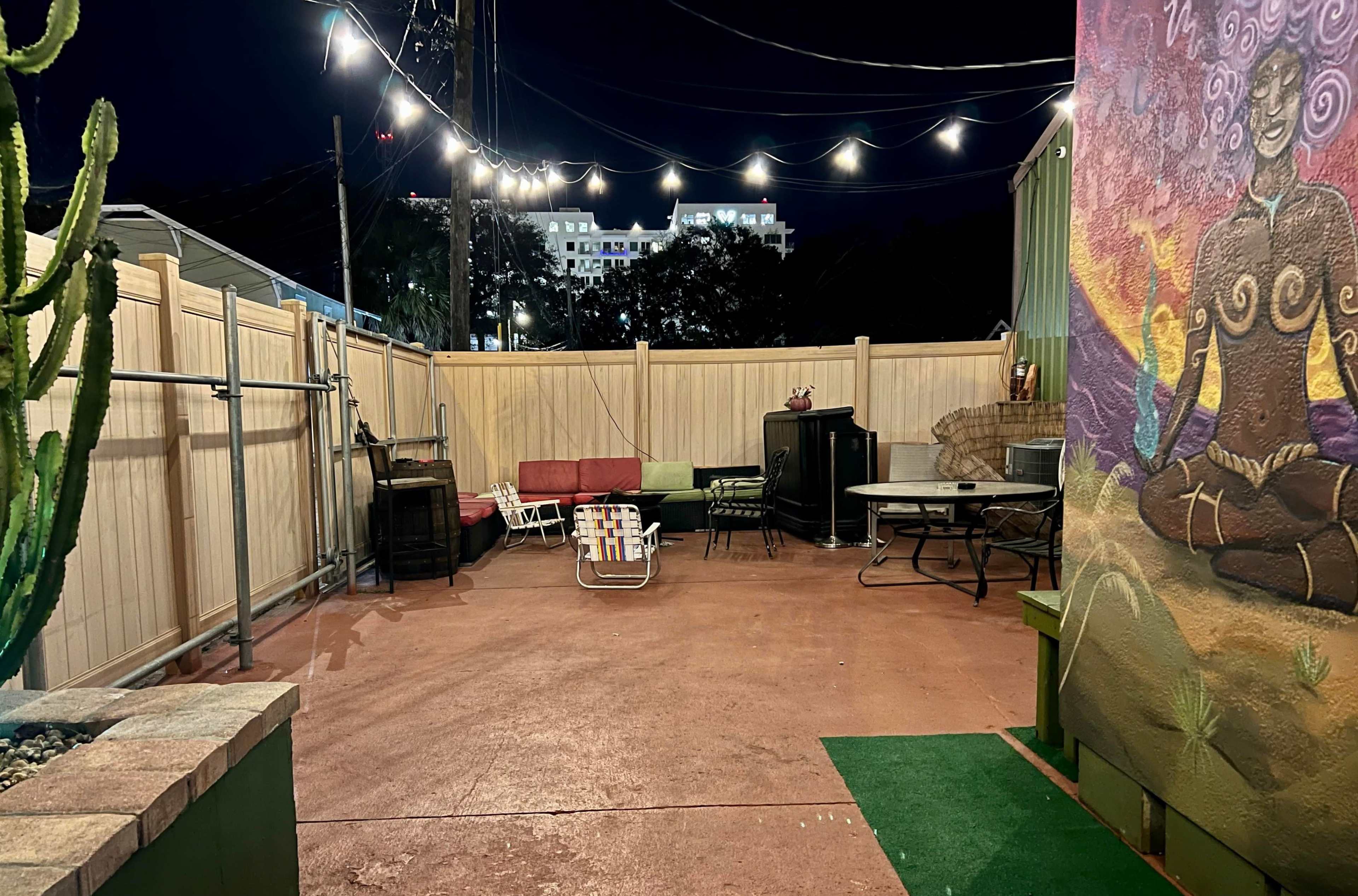 An outdoor patio area at night, featuring a seating arrangement with a red sofa, a folding chair, a dining set, and a wall mural.