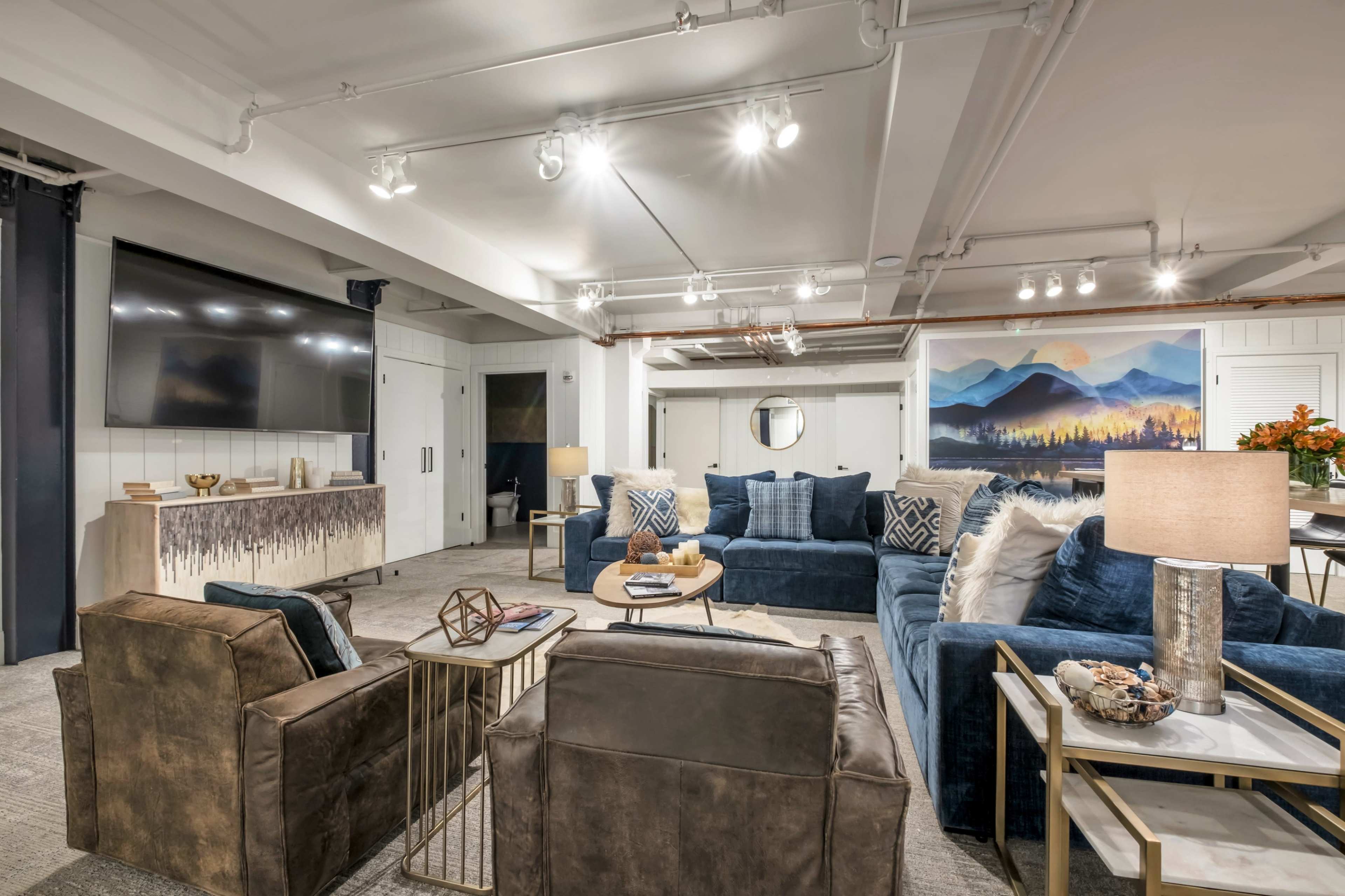 A modern lounge area features blue sofas, leather chairs, a wooden coffee table, and a wall mural depicting mountains.