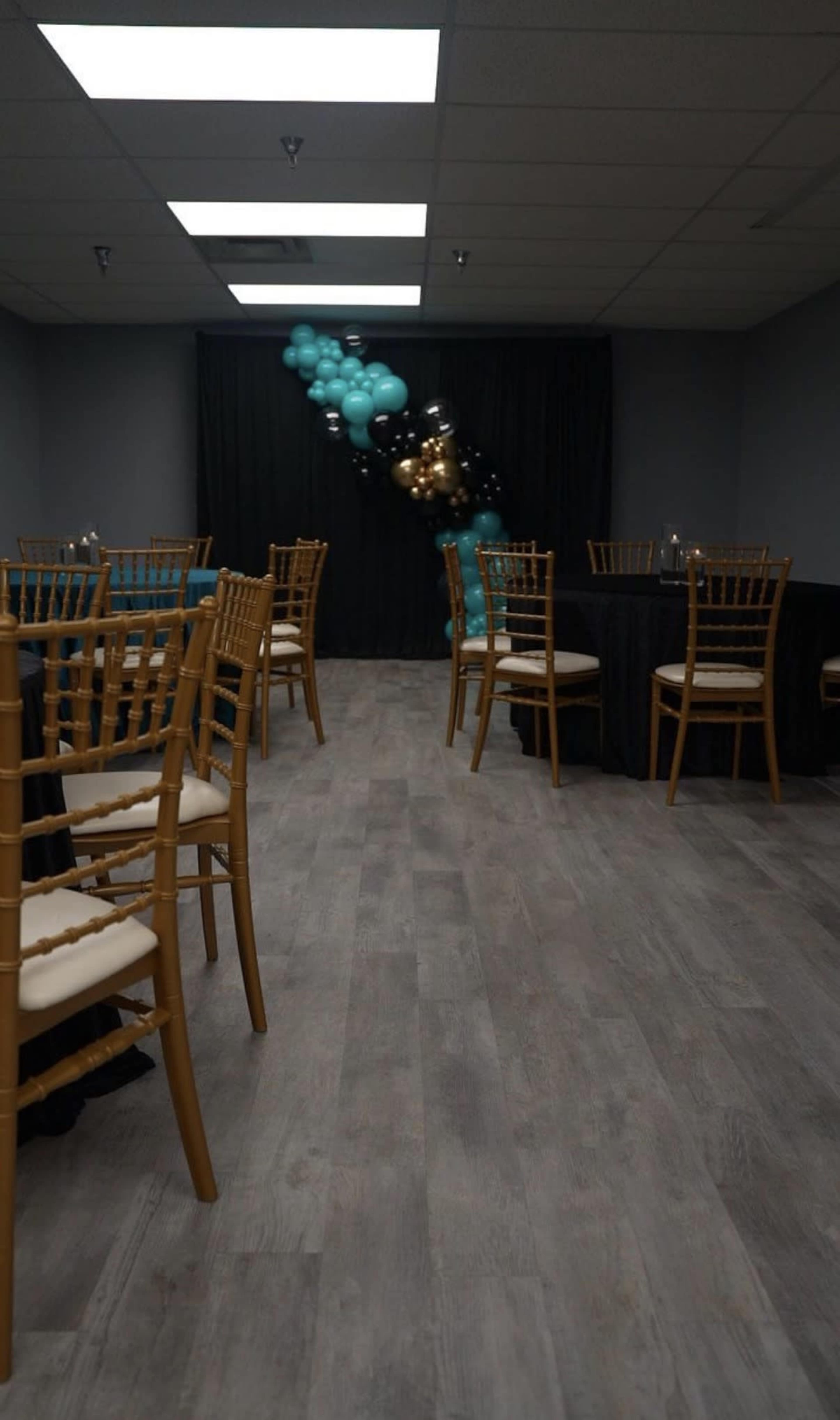 A decorated event space features wooden chairs and tables with black and teal tablecloths, along with a backdrop of balloons.