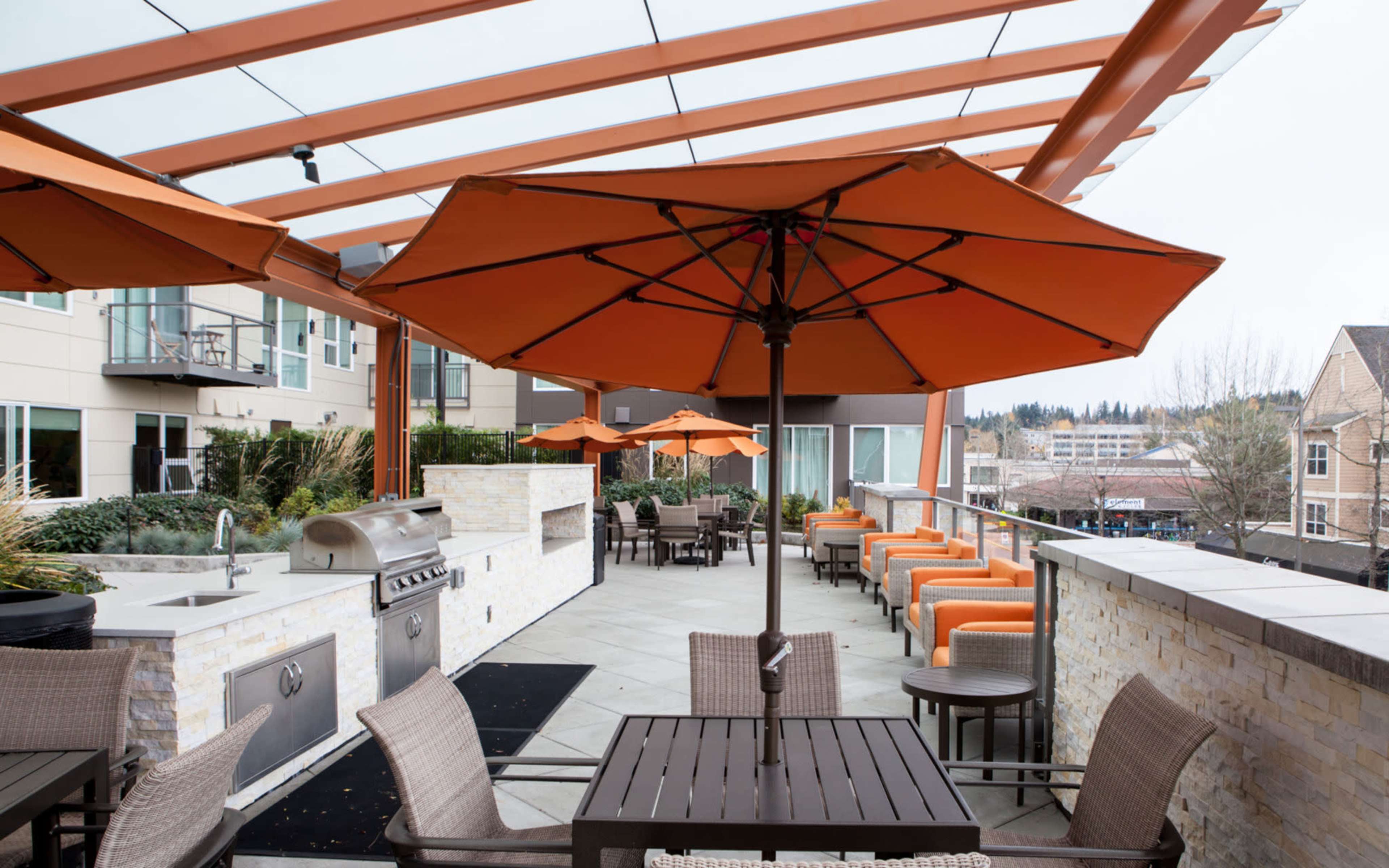 A rooftop terrace features orange umbrellas, patio seating, and a grill area surrounded by modern apartment buildings.