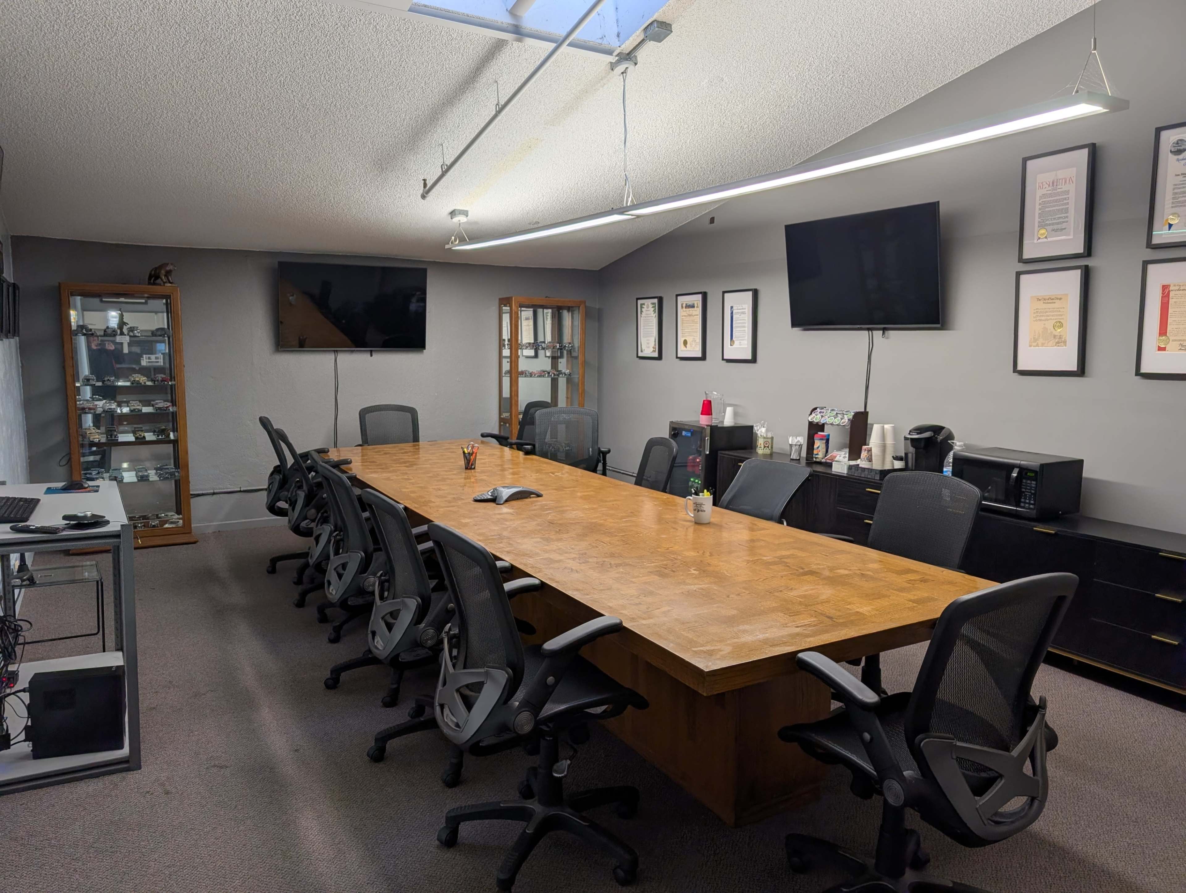 A conference room features a long wooden table surrounded by ergonomic chairs, with two wall-mounted screens and a cabinet displaying various items.