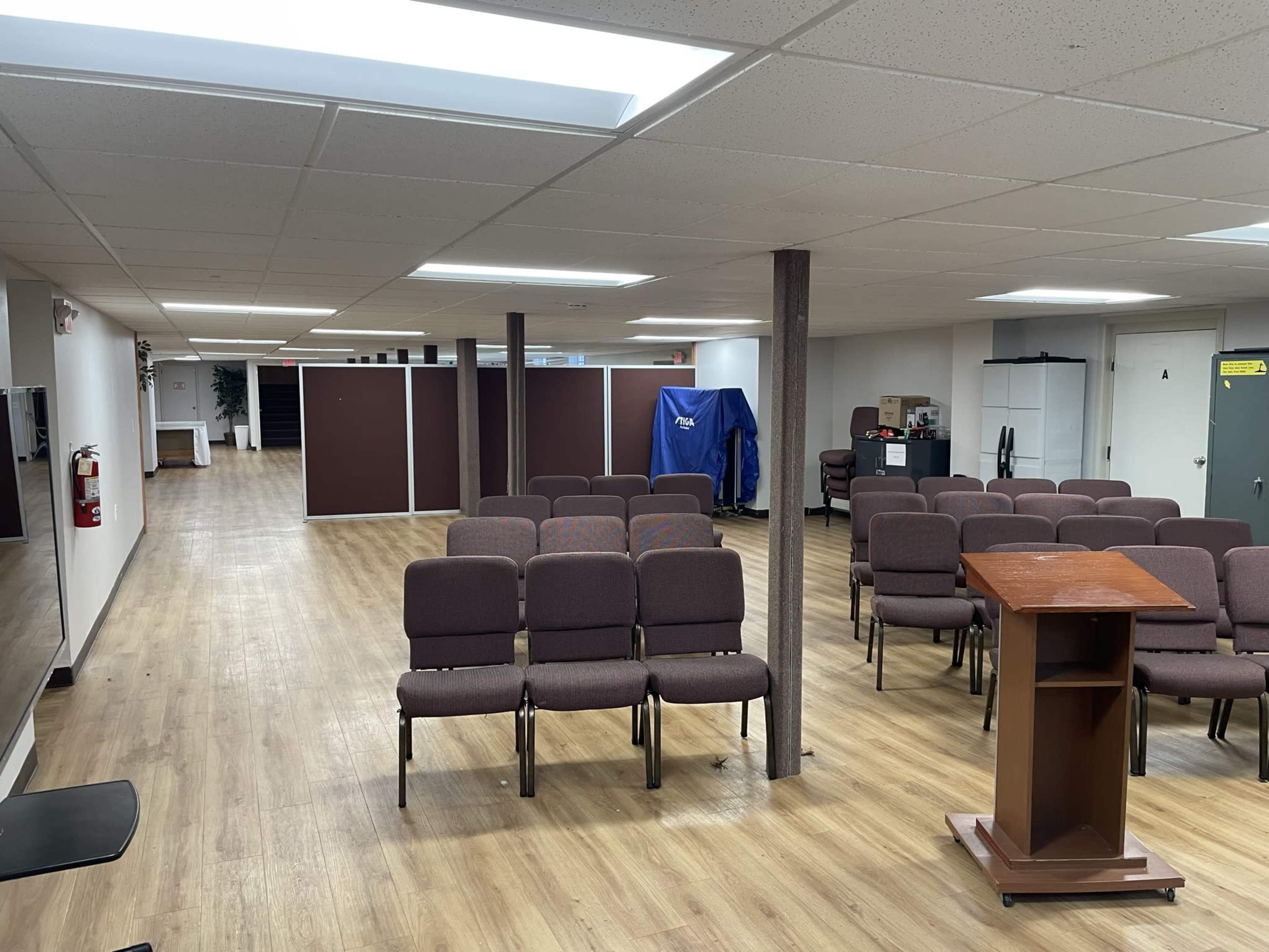 A spacious room with rows of chairs facing a wooden podium, partitioned areas, and light-colored flooring.