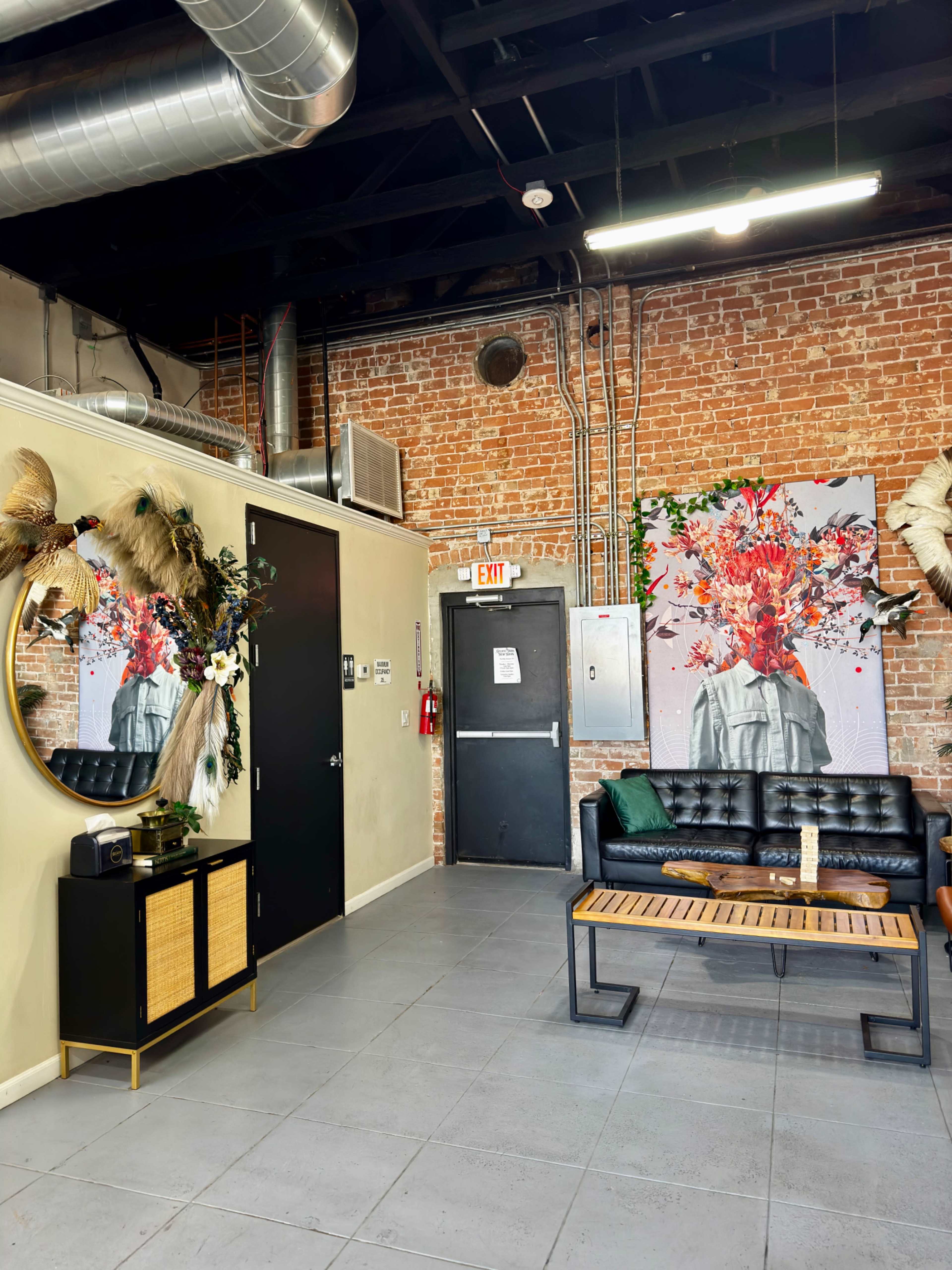 The image depicts an interior space with exposed brick walls, a black sofa, a round mirror with decorative flowers, and vibrant artwork hanging on the wall.