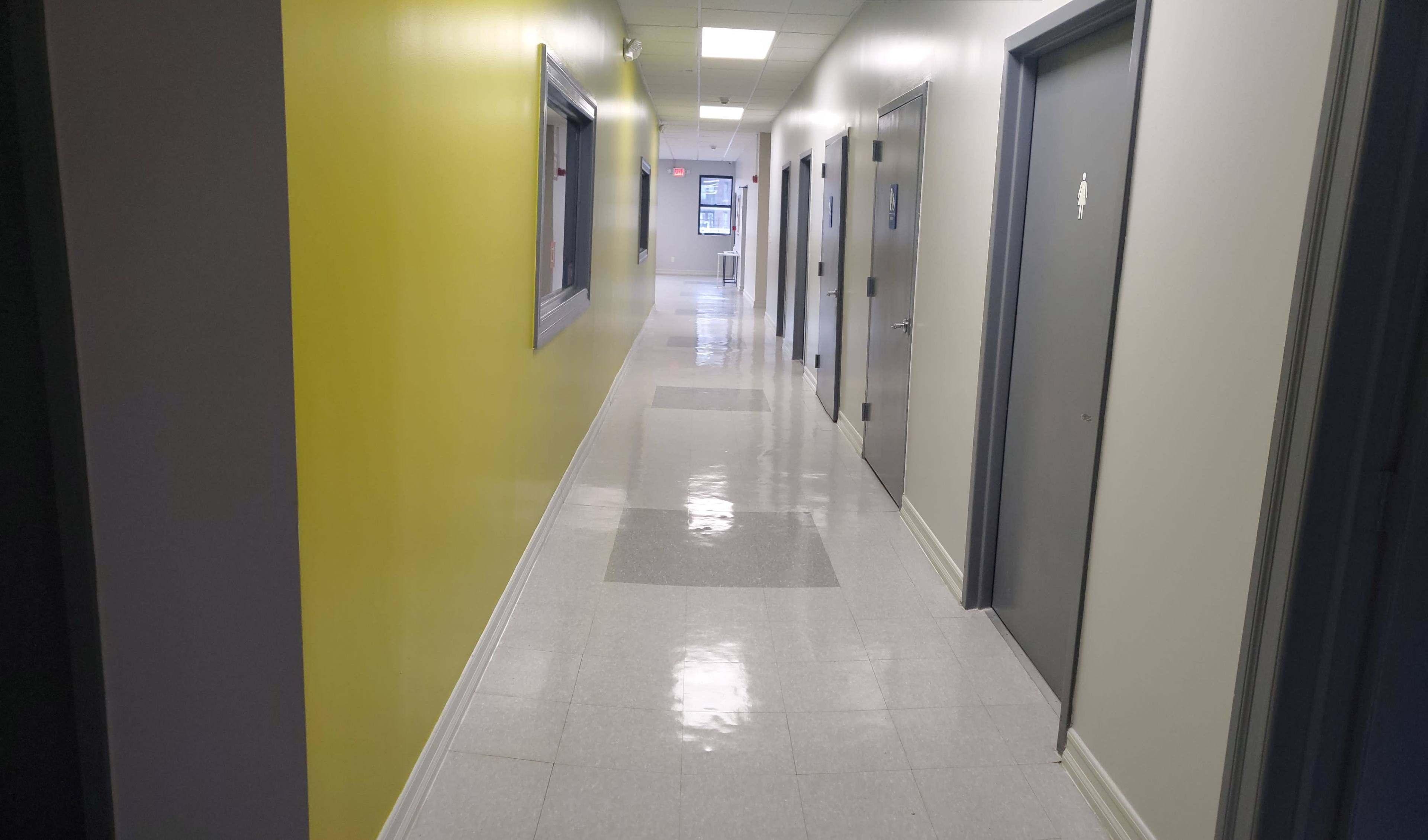 A corridor with yellow and neutral walls, featuring several doors and shiny tile flooring, leads to a window at the far end.