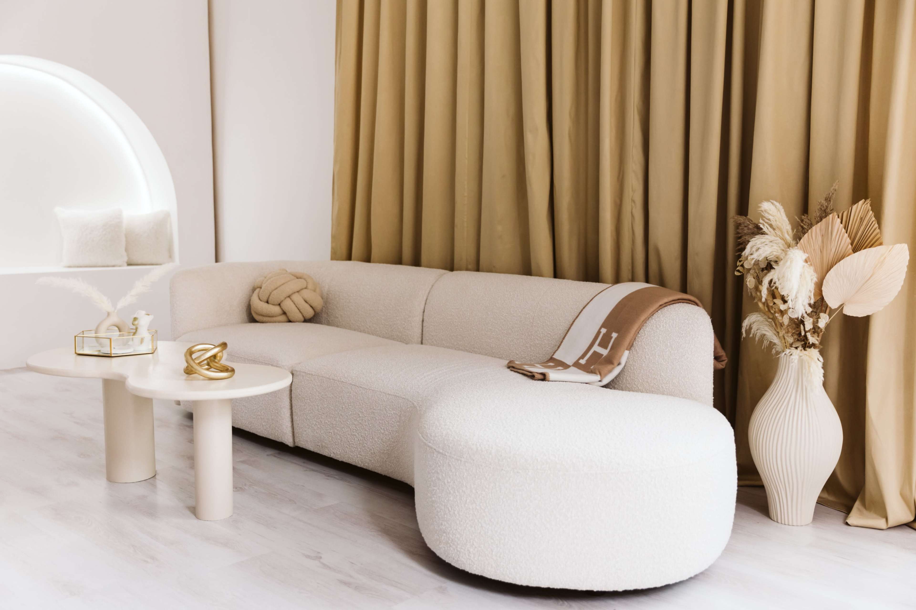 A modern living room featuring a white, curved sectional sofa, a small round coffee table, and a decorative vase with dried plants, all against a backdrop of soft beige curtains.