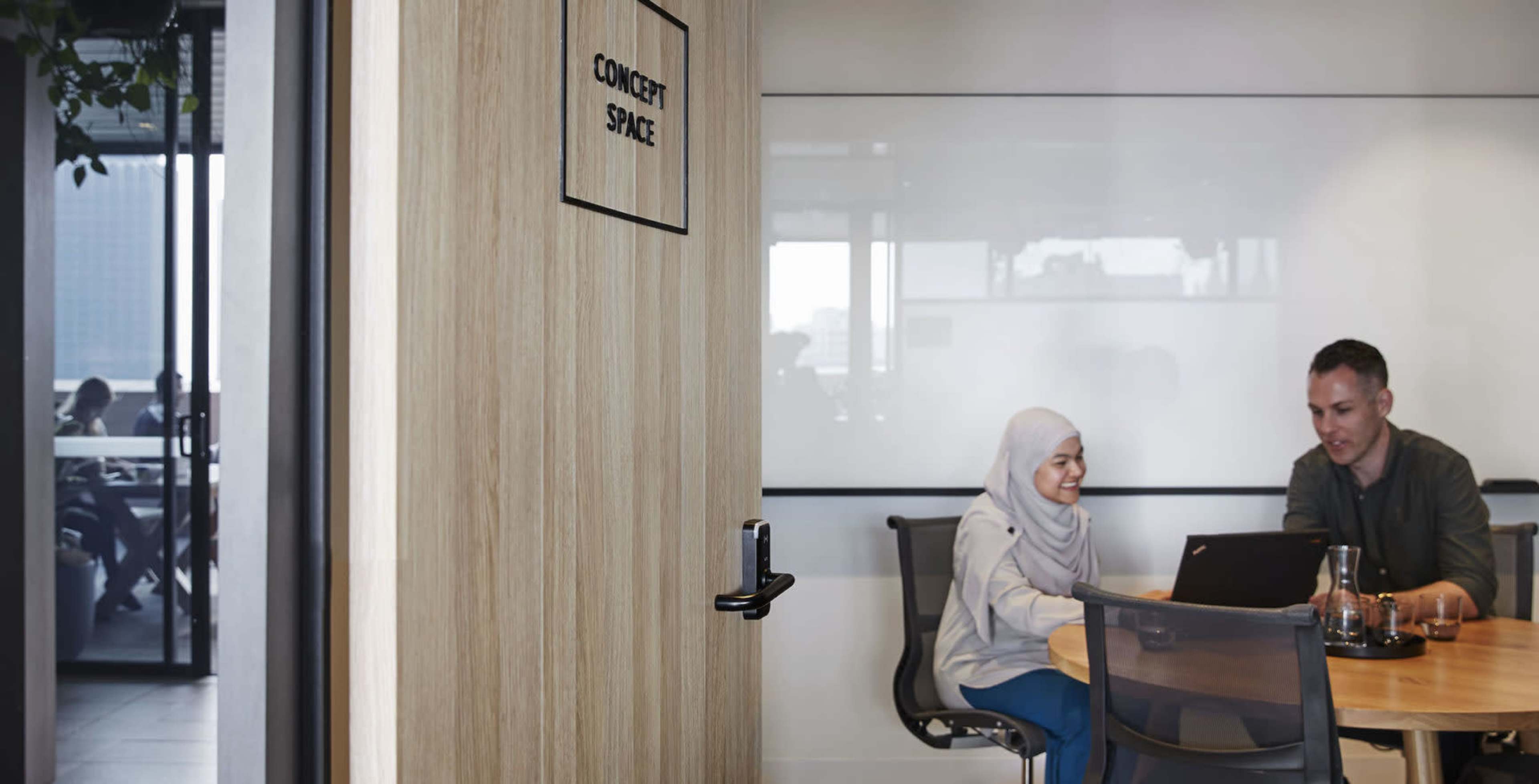 A person wearing a hijab and another individual are seated at a table in a room labeled "CONCEPT SPACE," working on a laptop.