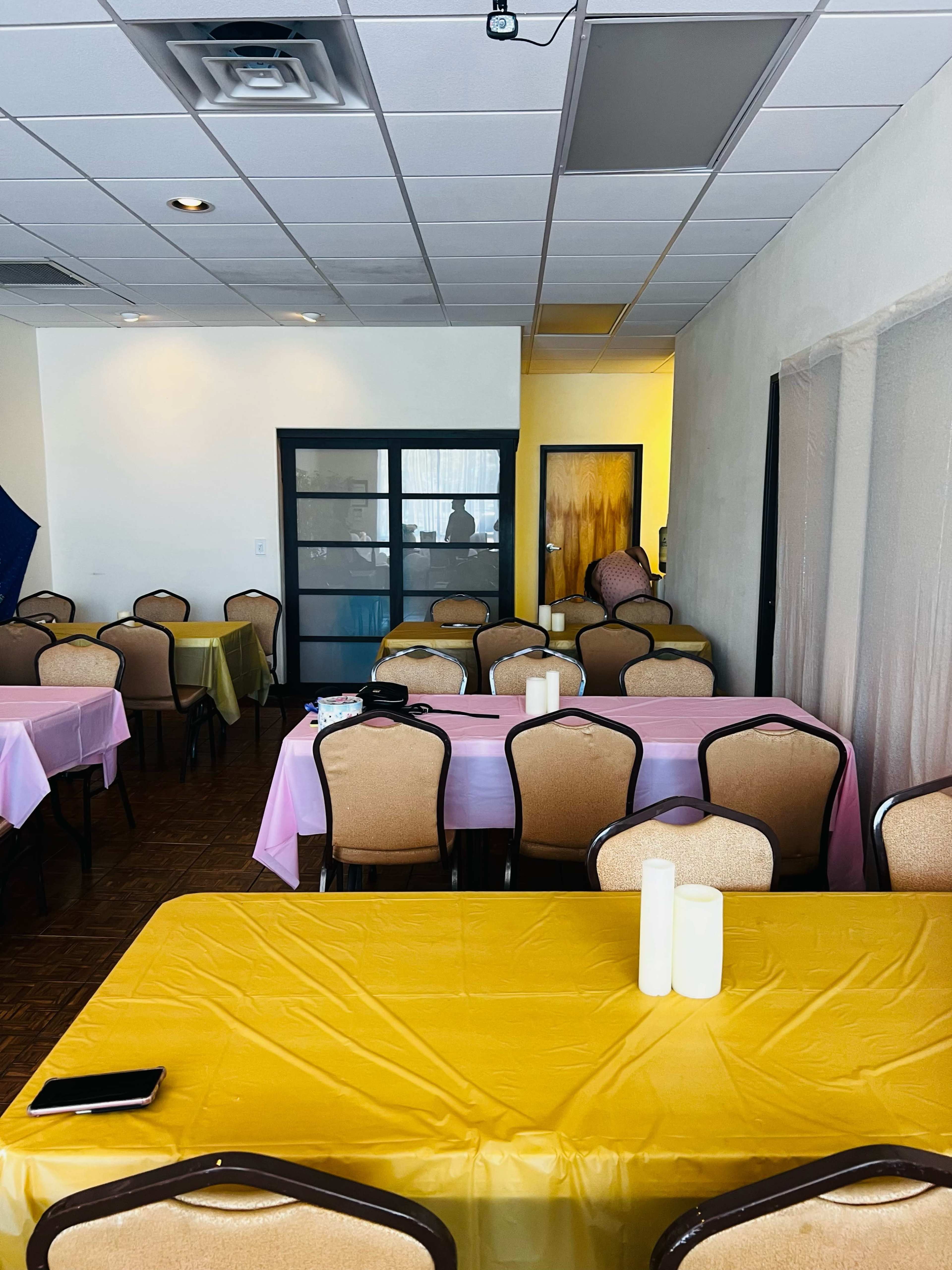 A dining area with several tables covered in yellow and pink tablecloths, some chairs arranged around them, and a light-colored wall in the background.