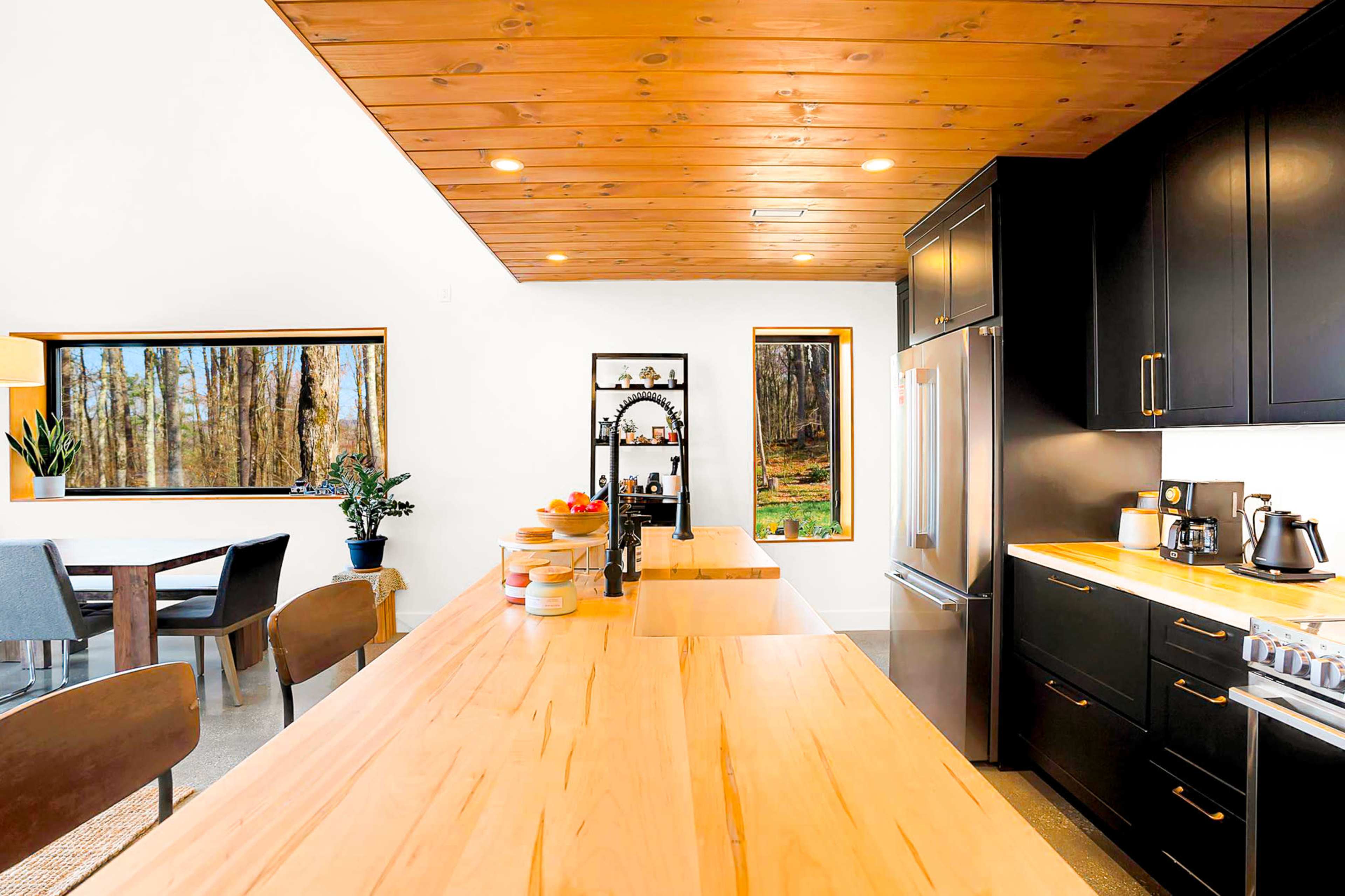 A modern kitchen features a large wooden island, black cabinetry, and windows that provide a view of a wooded area outside.