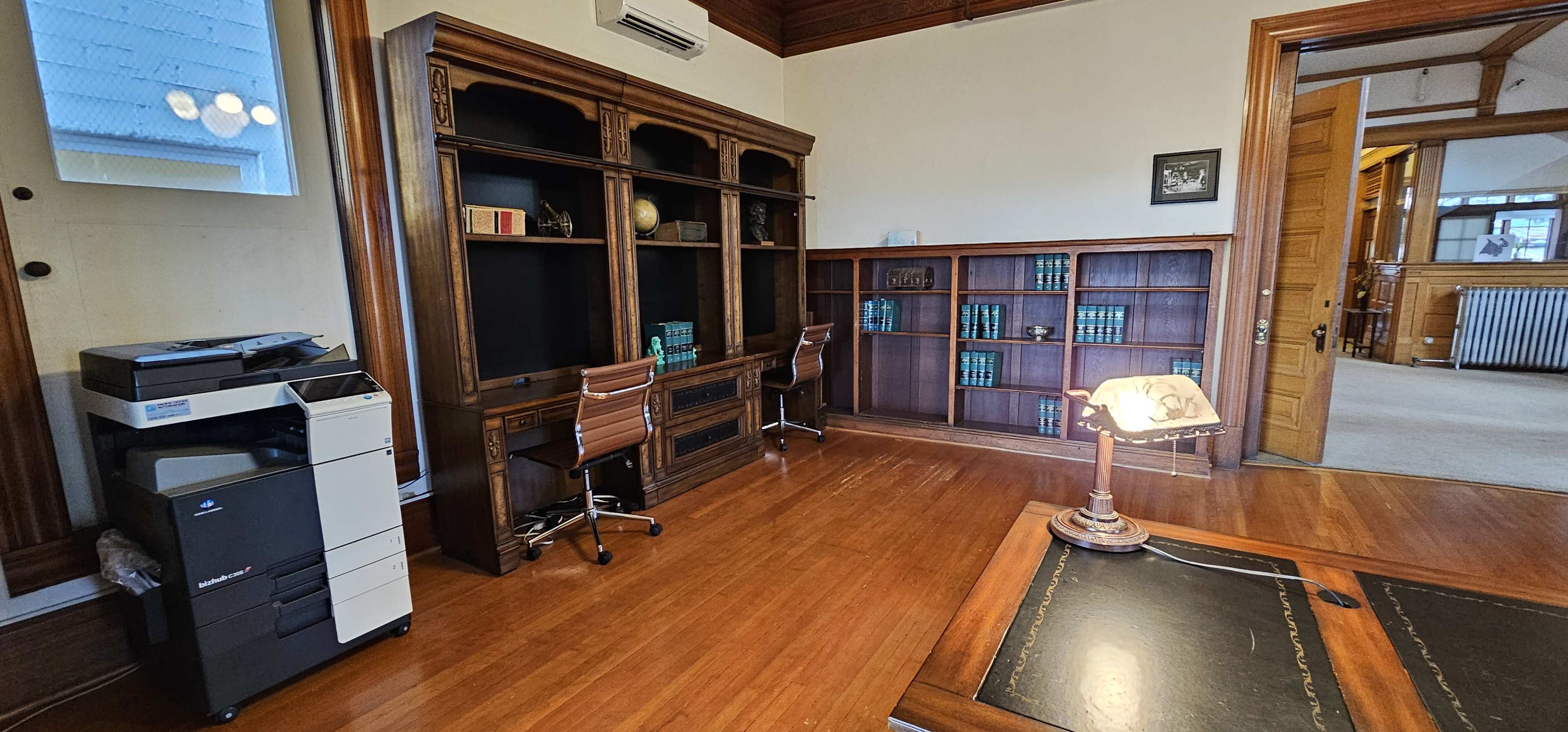 A spacious office with wooden bookshelves, a printer, and a desk lamp, featuring a wooden floor and an adjoining room.