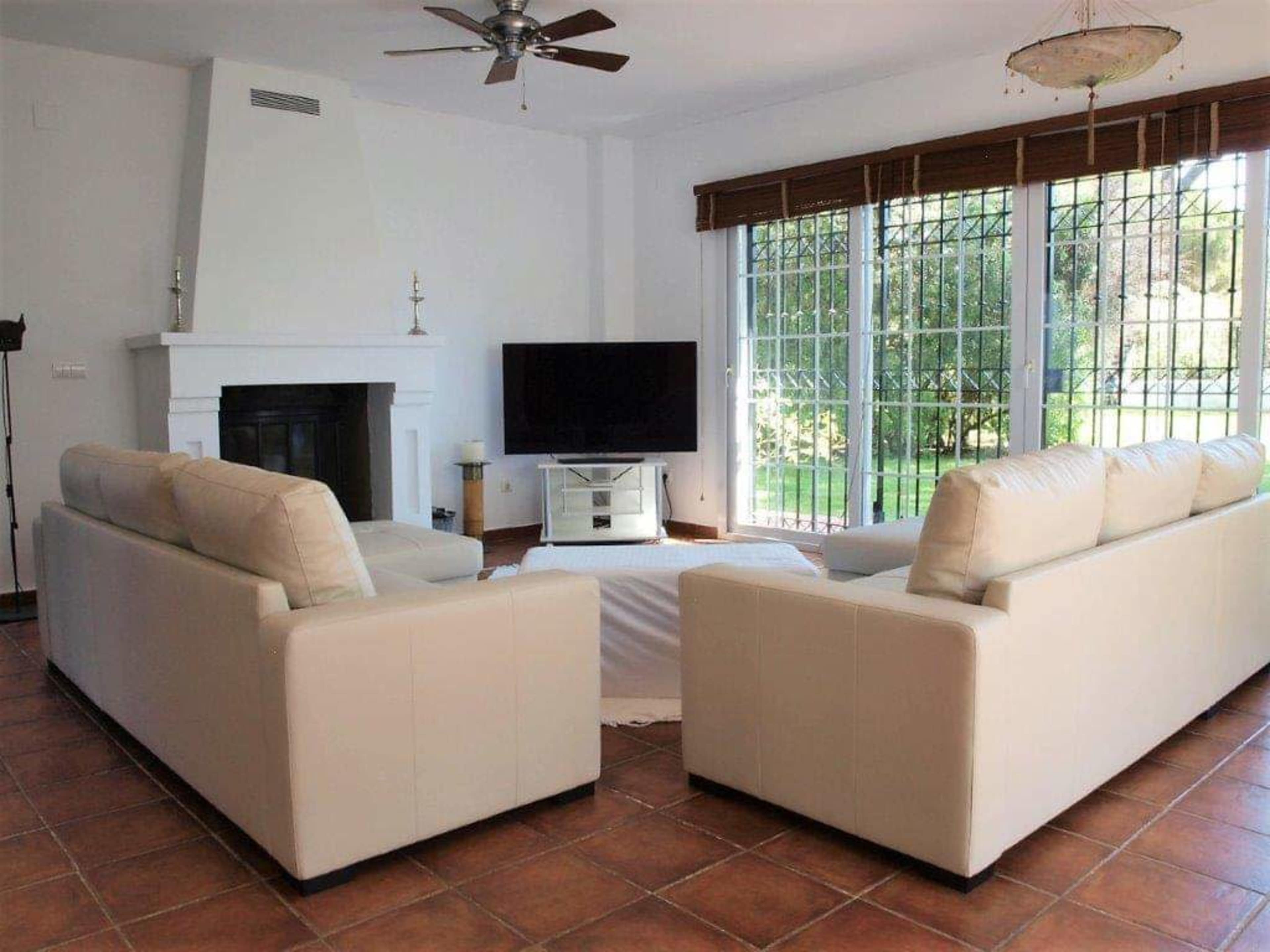 The image shows a bright living room with two white sofas facing each other, a television mounted on the wall, and large windows overlooking a garden.