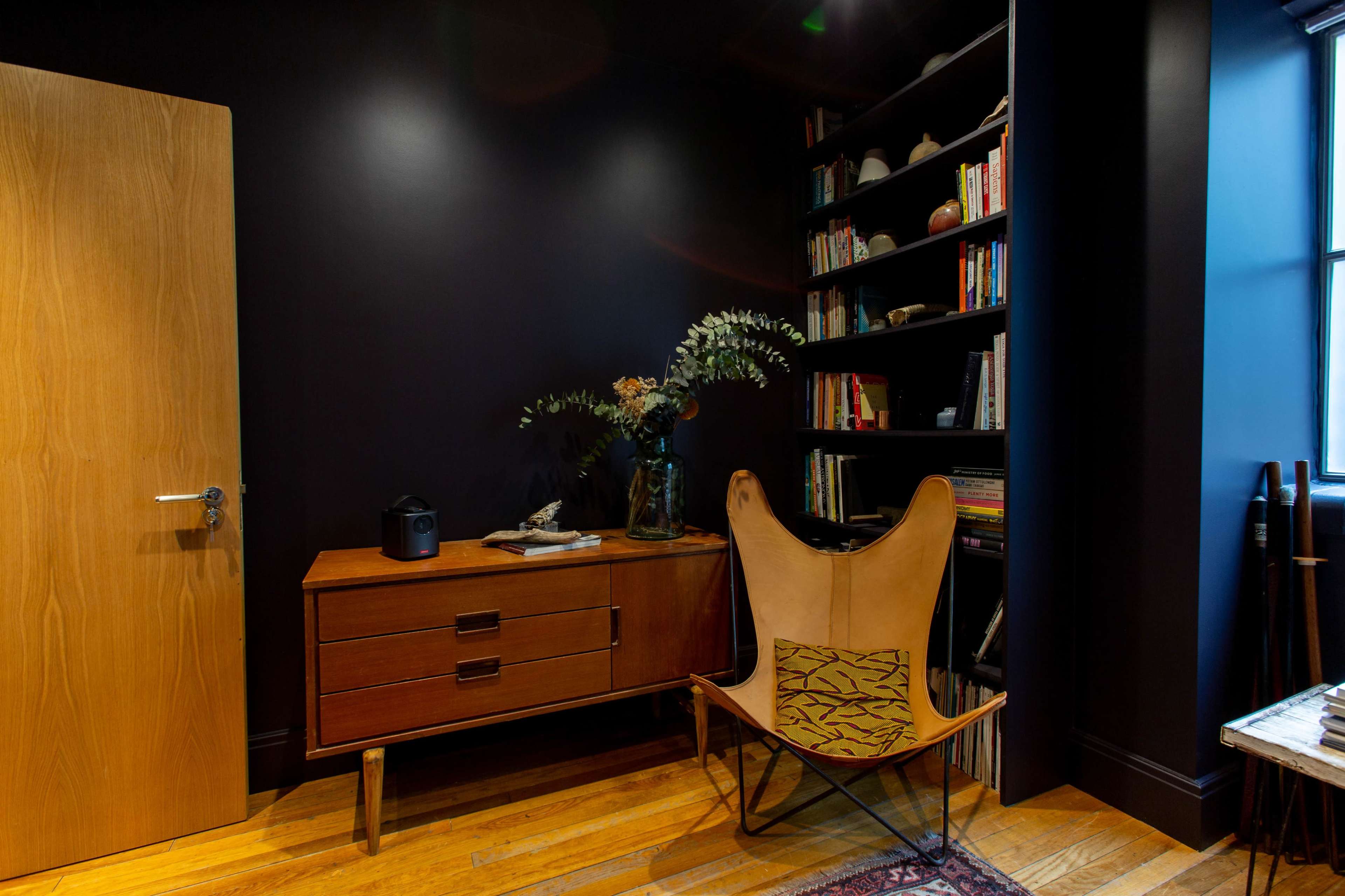 A cozy corner of a room features a wooden cabinet, a potted plant in a vase, a butterfly chair, and a bookshelf filled with books.
