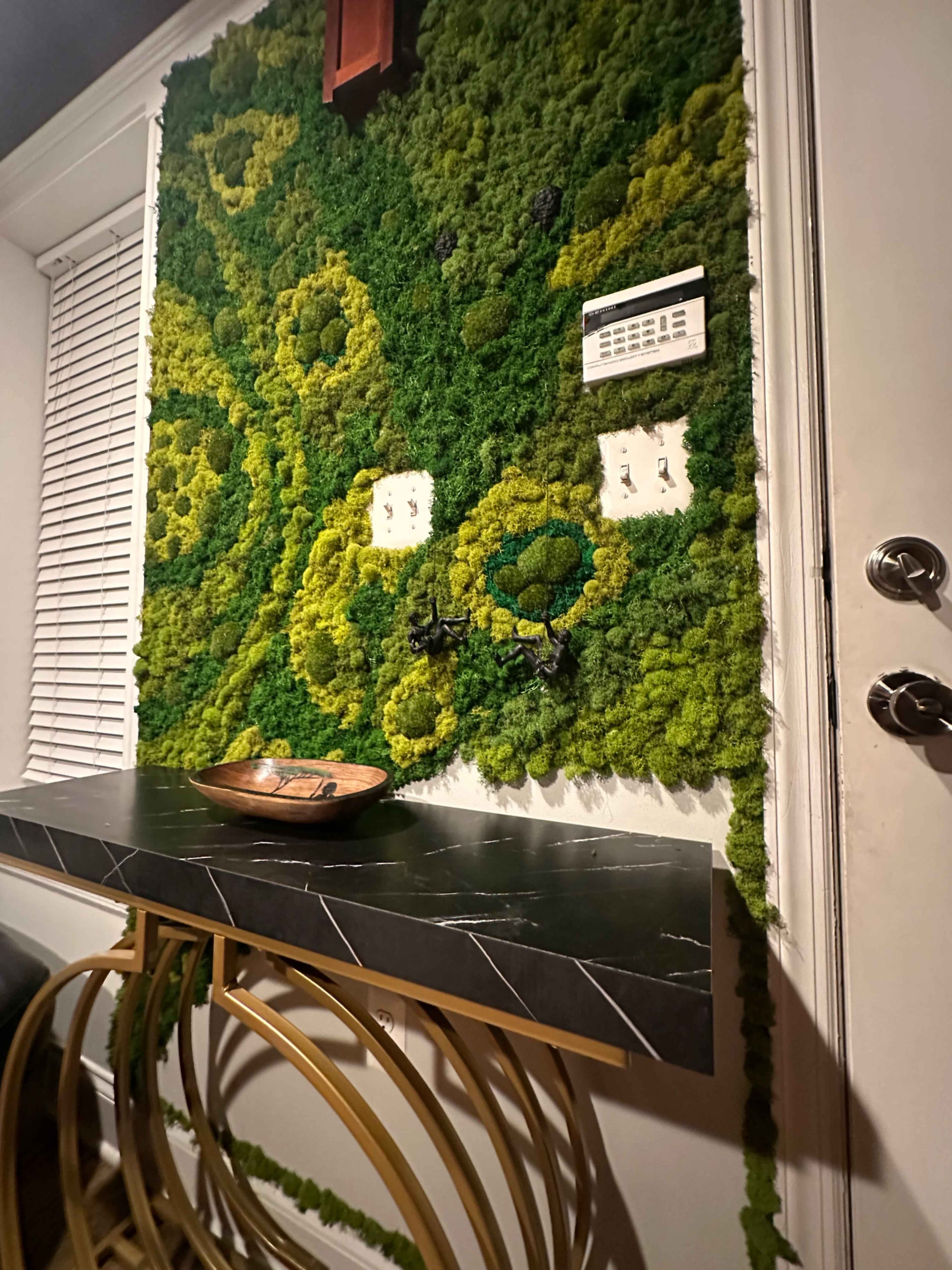 A textured green moss wall decorates a room behind a dark marble-top console table with decorative items.