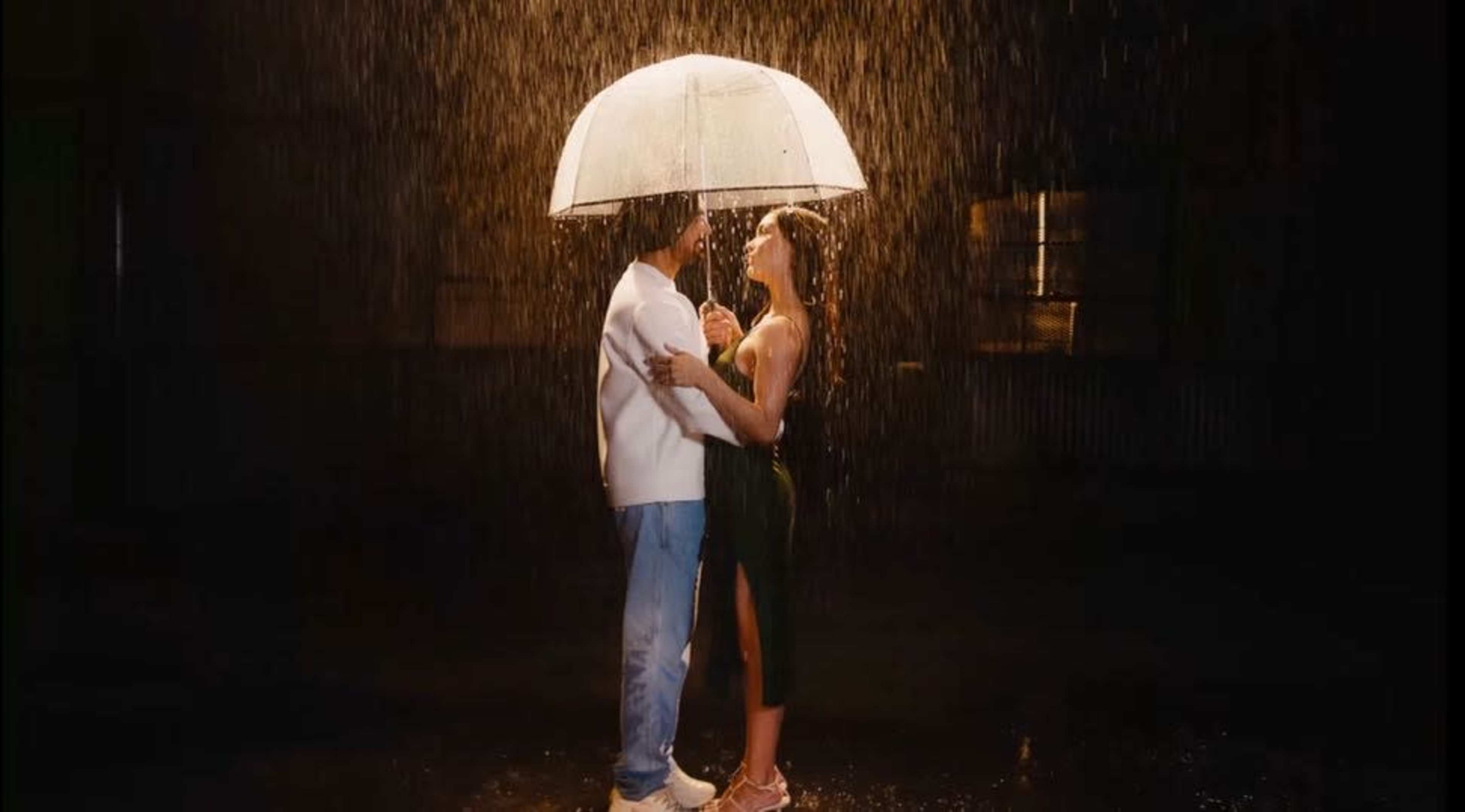 A couple stands under a large umbrella, embracing in the rain against a dimly lit backdrop.