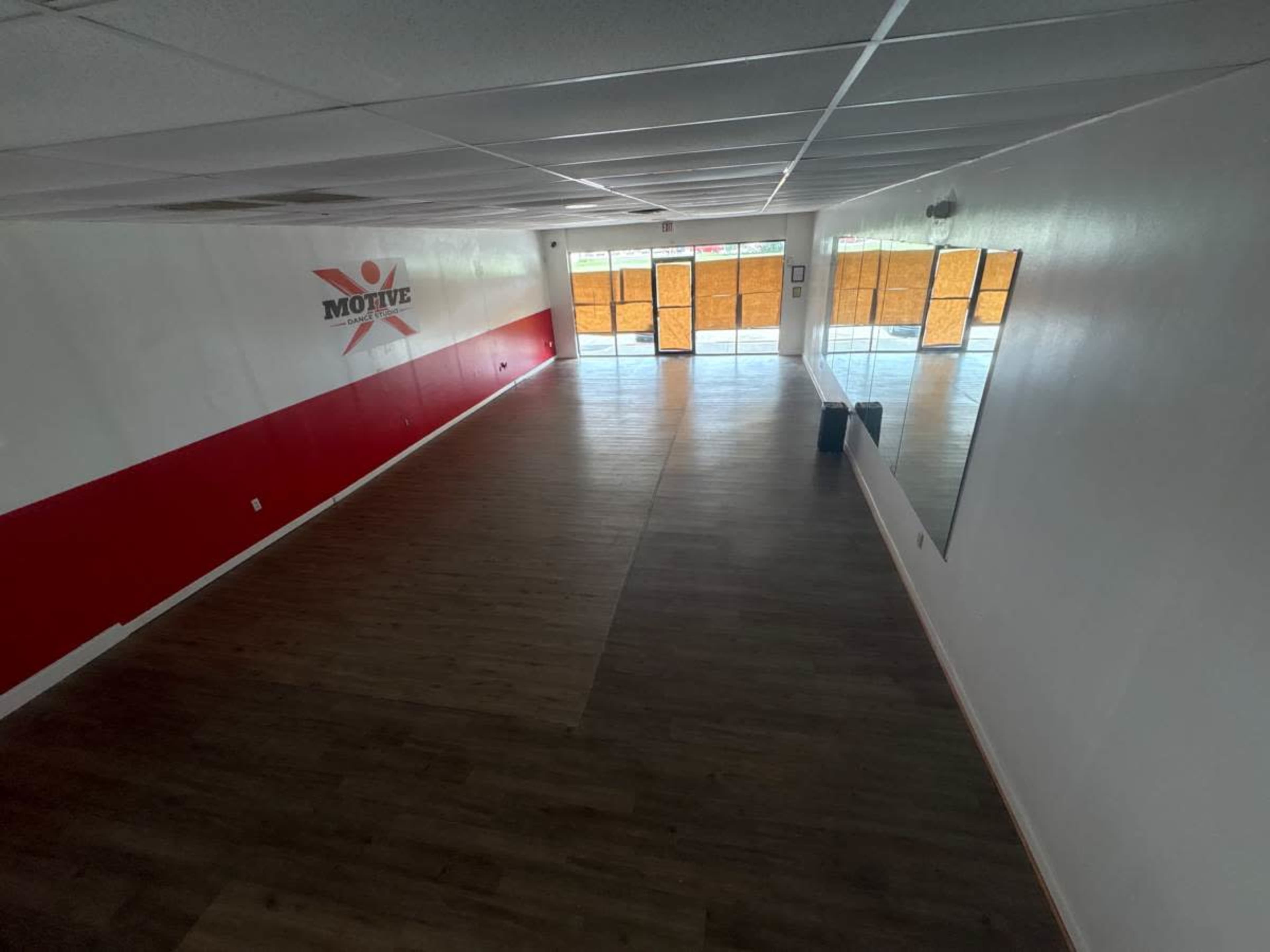 The image shows an empty, well-lit interior space with wooden flooring, a mirrored wall, and large windows partially covered by an orange shade.
