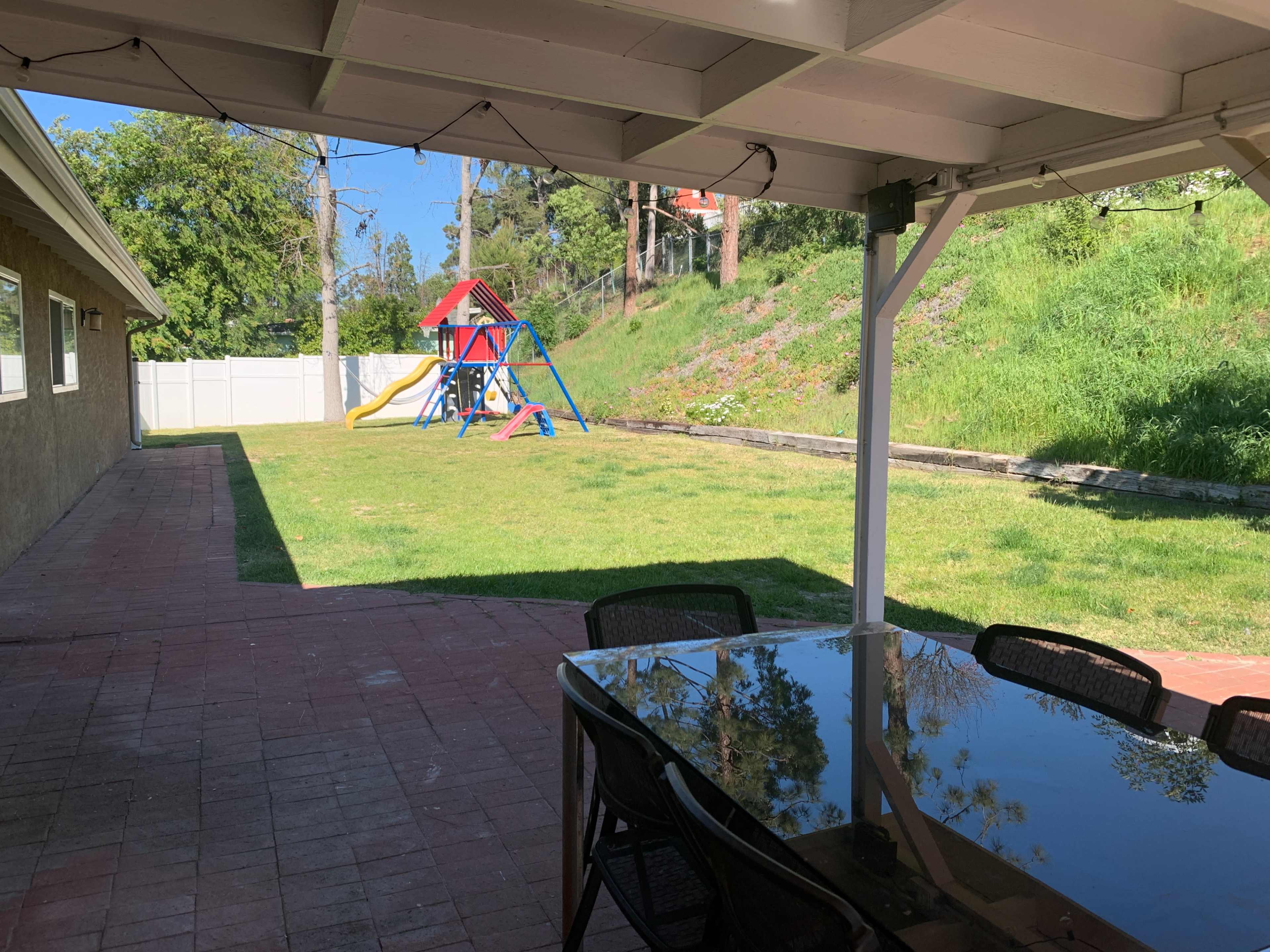 A backyard features a playground with a slide and swings, viewed from a patio with a black table and chairs.
