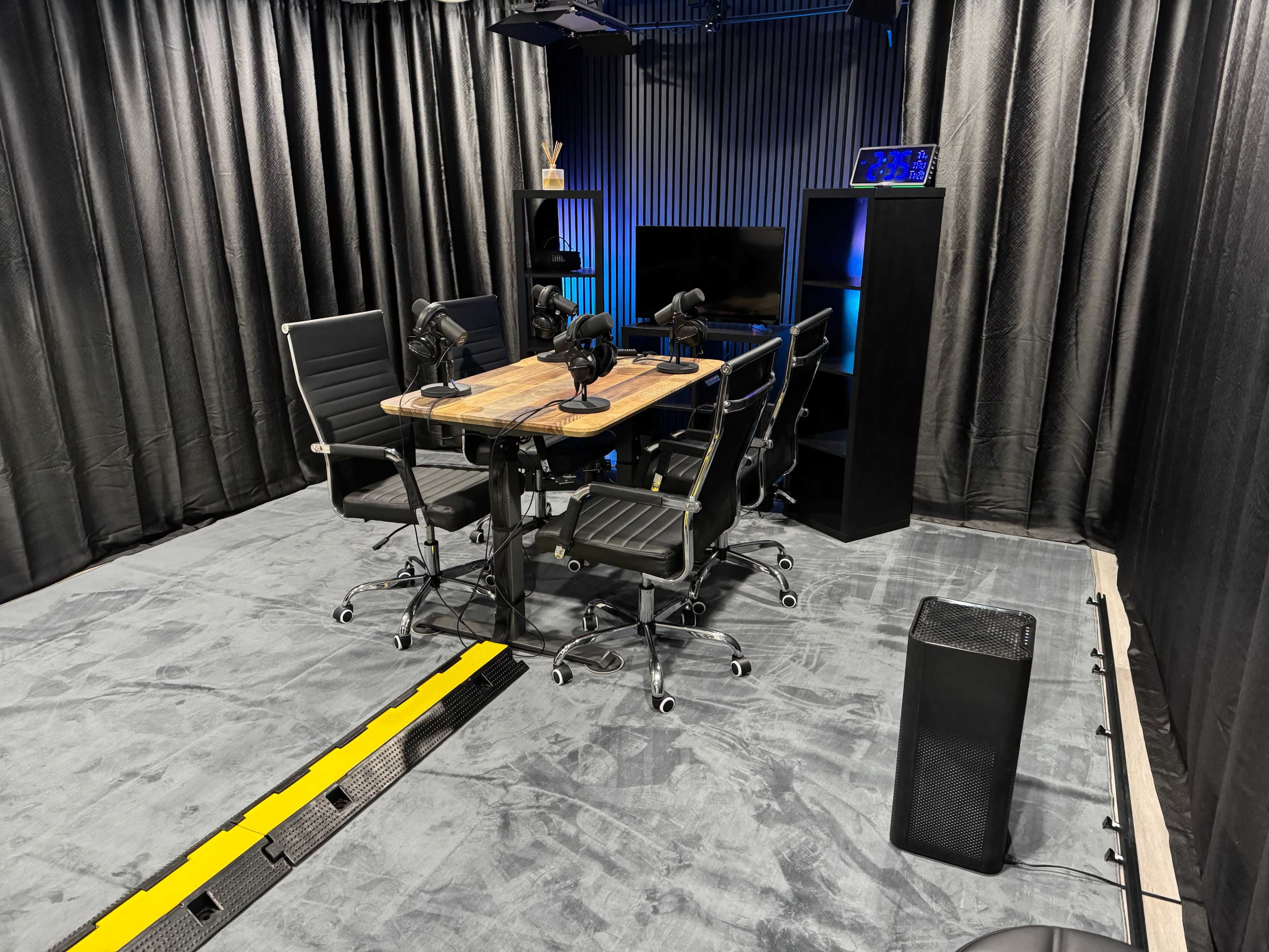 The image shows a recording studio setup with a wooden table surrounded by four black chairs and microphones, along with a dark backdrop and a television on a shelf.