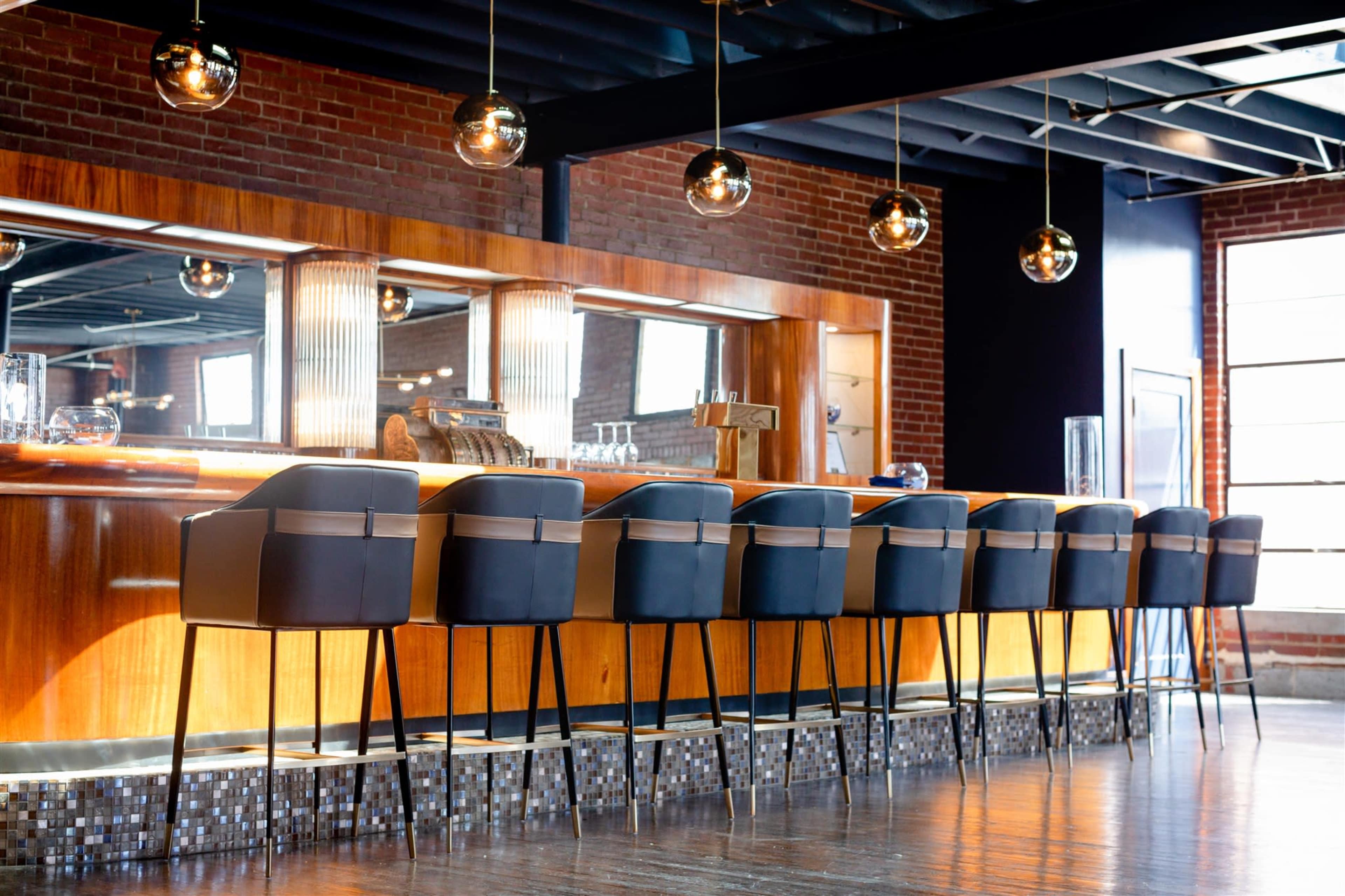 A modern bar features a row of black upholstered stools facing a polished wooden counter, illuminated by pendant lights, against a backdrop of brick walls and large windows.