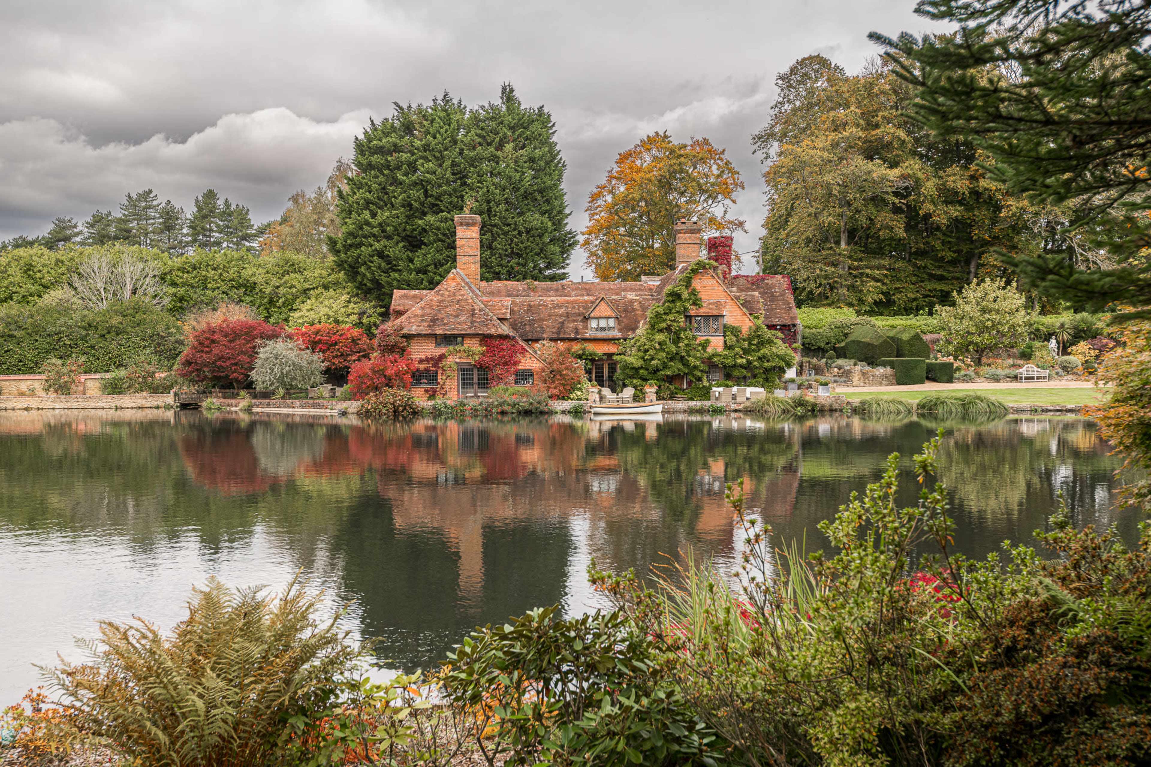 A quaint cottage with a red-roofed exterior is situated beside a serene lake, surrounded by lush greenery and autumn-colored trees.