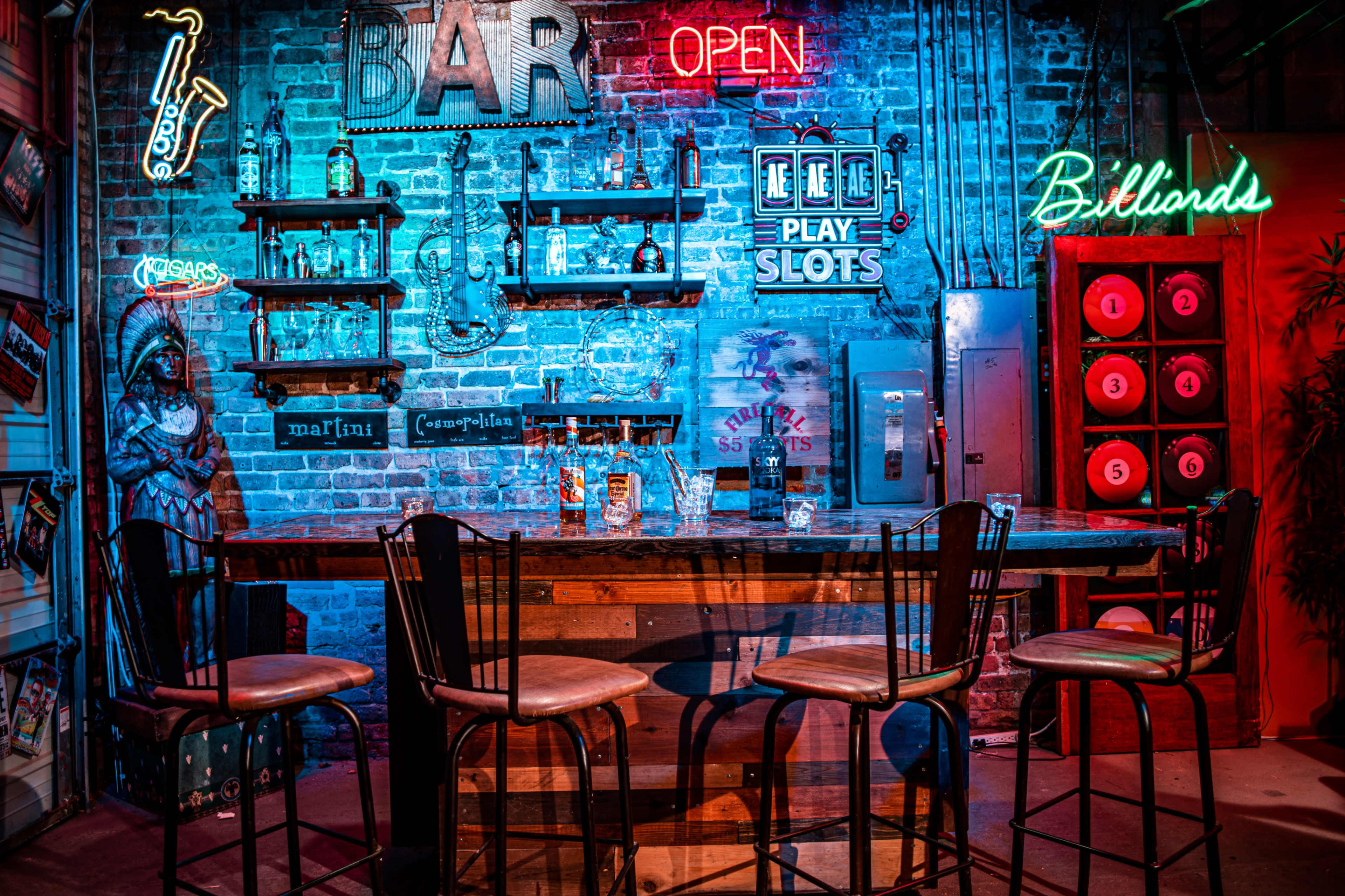 A dimly lit bar scene features a brick wall adorned with neon signs, shelves of liquor bottles, and wooden bar stools arranged around a large wooden bar.